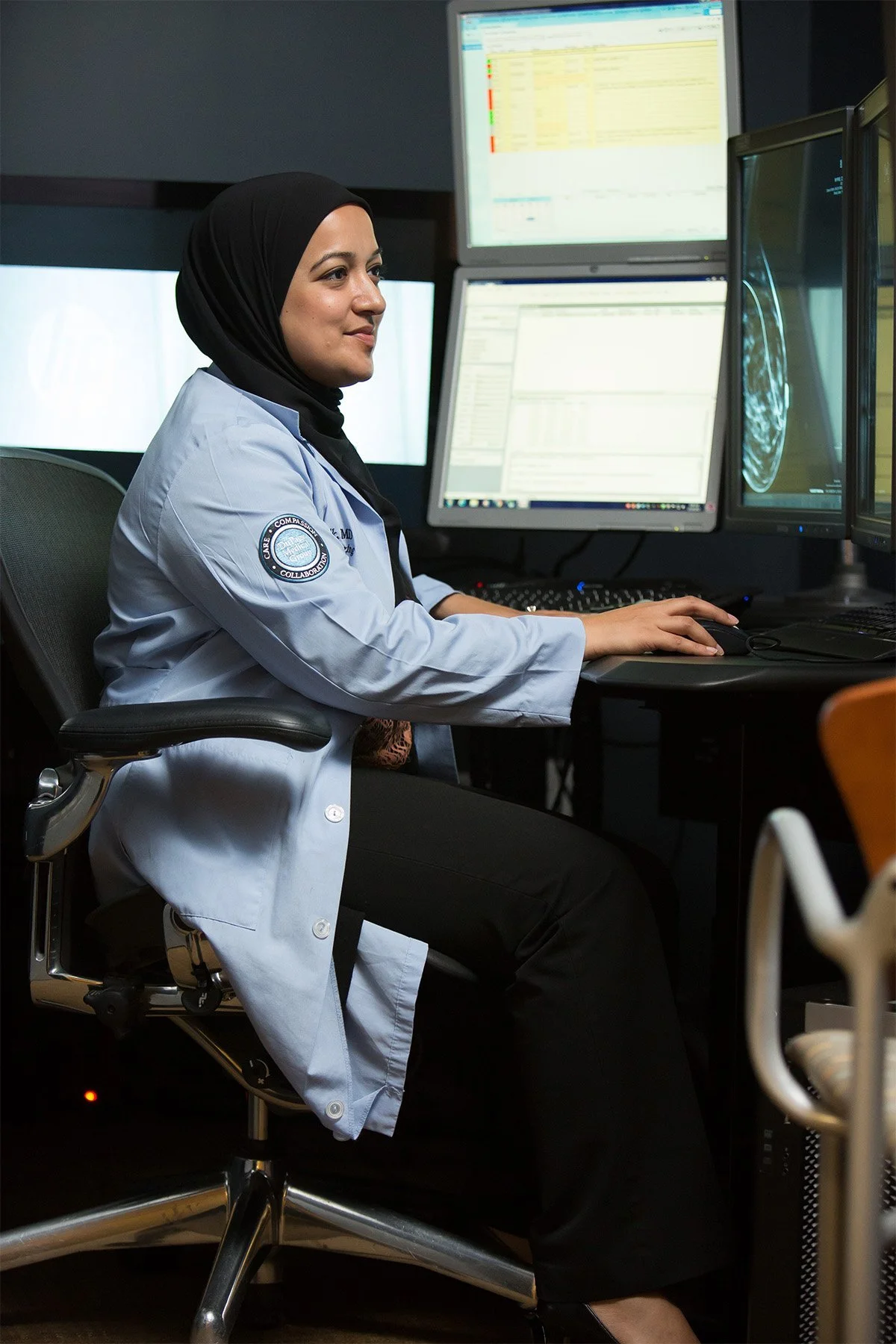 A woman in a white medical coat with a patch on her arm, wearing a black hijab, is seated at a desk with multiple computer monitors displaying medical images and data, in a clinical or hospital setting.