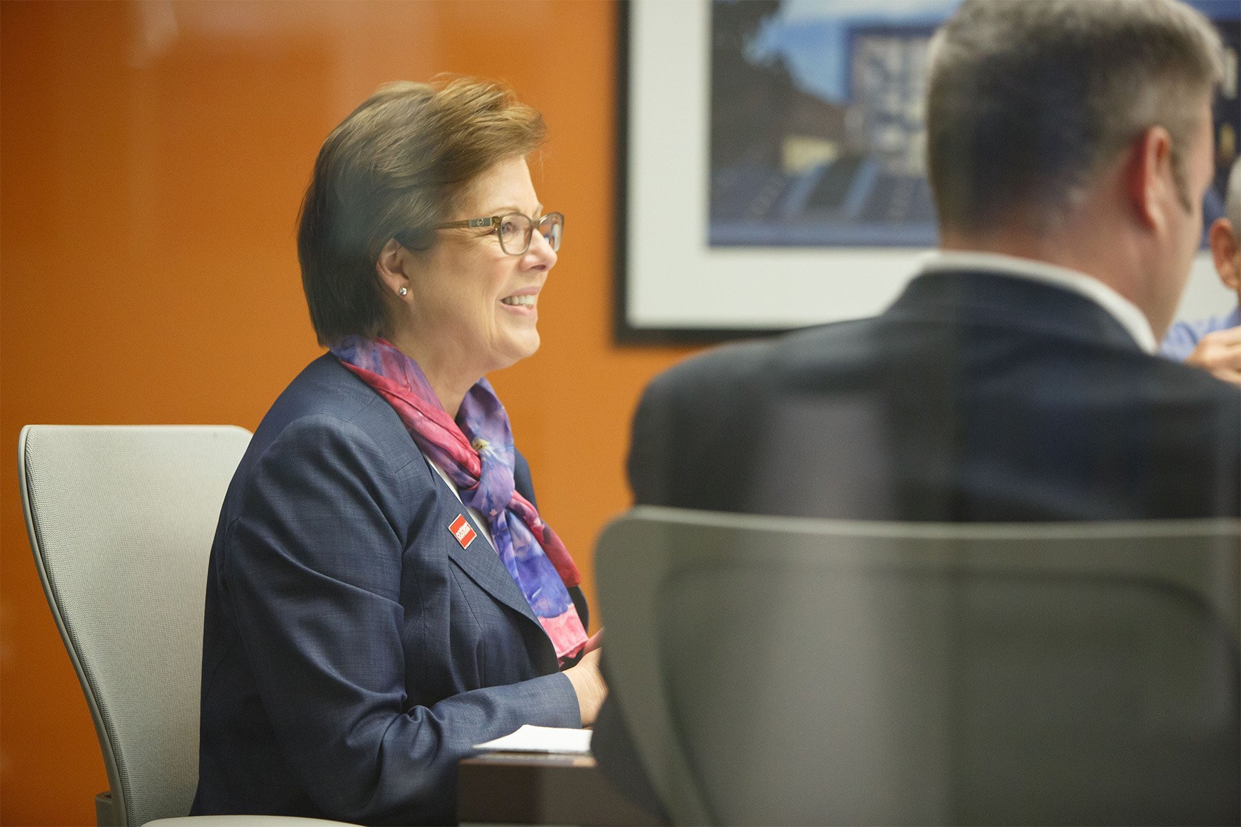 A woman with glasses and short hair smiling during a meeting, wearing a navy blazer with a red, white, and purple scarf, sitting at a table with a white chair against an orange wall. A man with short hair in a dark suit is sitting beside her, facing 