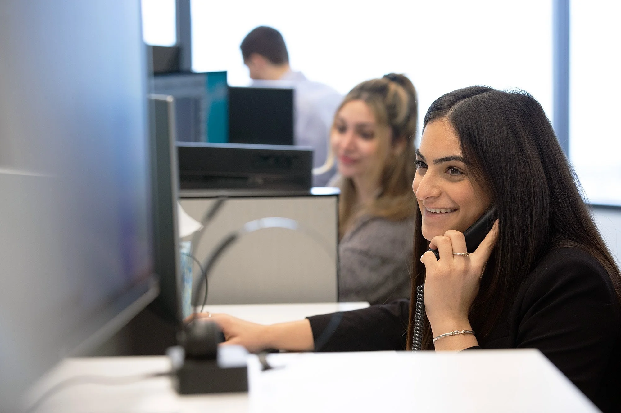 A woman with long dark hair smiling while talking on the phone at her desk in an office, with two other colleagues working at their desks in the background.
