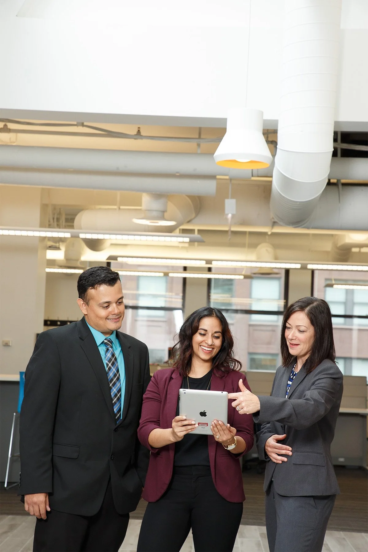 Three business professionals, two women and one man, standing together in an office, looking at a tablet device held by the woman in the middle, with smiles and engaged expressions.