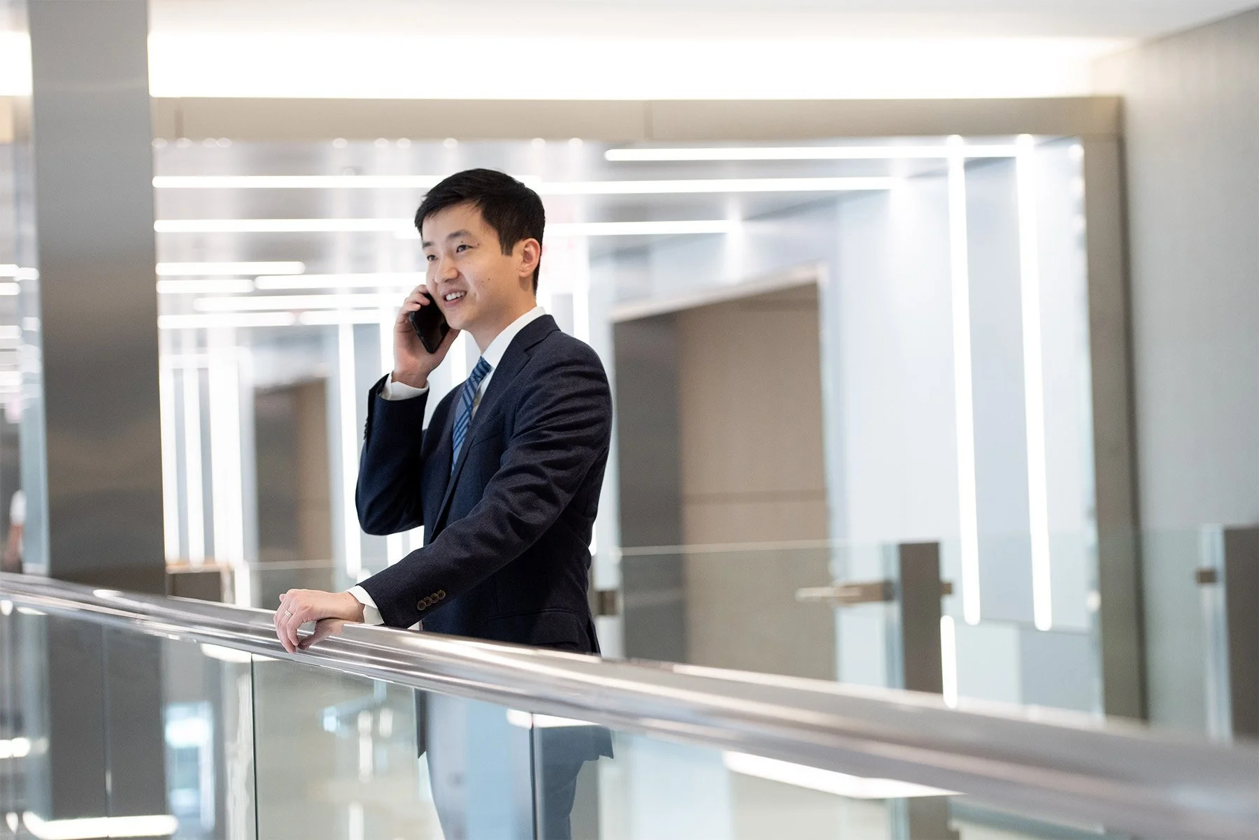 A young Asian man in a dark suit talking on a cellphone and standing at a railing in a modern, well-lit building with reflective surfaces and horizontal light fixtures.