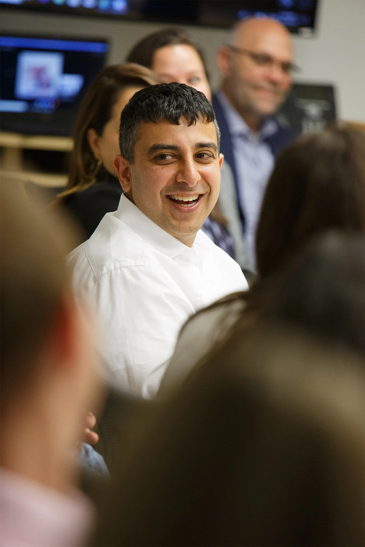 A man in a white shirt smiling during a meeting or seminar, with other adults seated around him in a conference room.