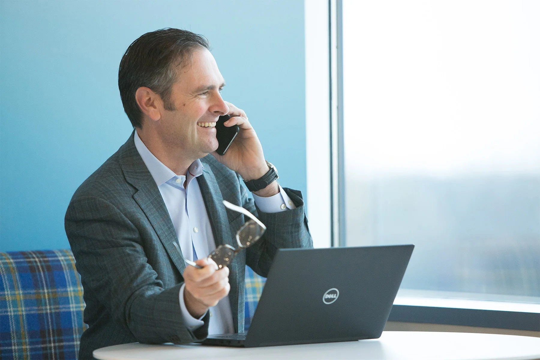 A man in a checkered blazer and white shirt, smiling while on a phone call, holding his glasses in one hand, with a Dell laptop open in front of him in a bright office setting.
