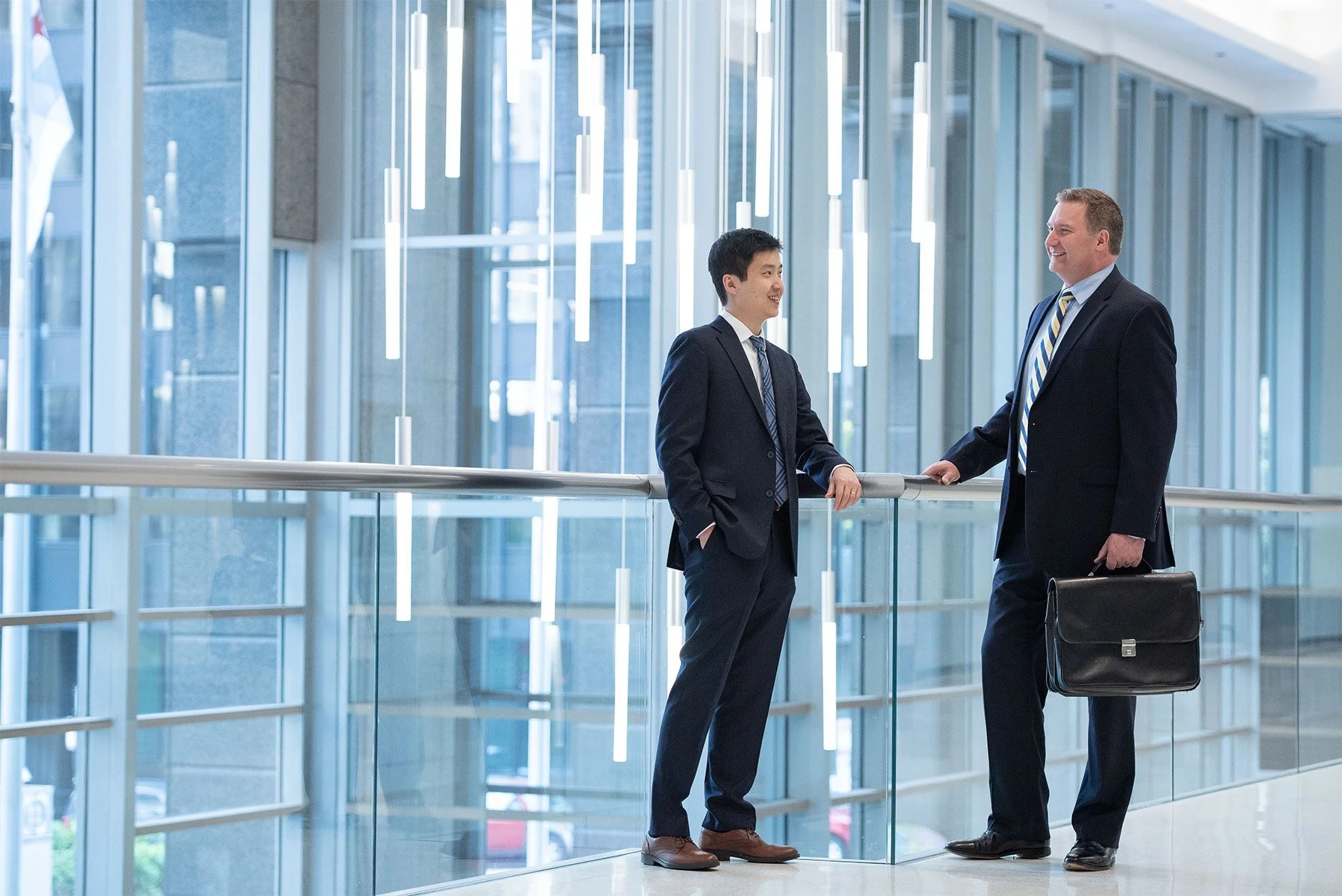 Two businessmen in suits having a conversation in a modern office building with large glass windows and hanging lights.