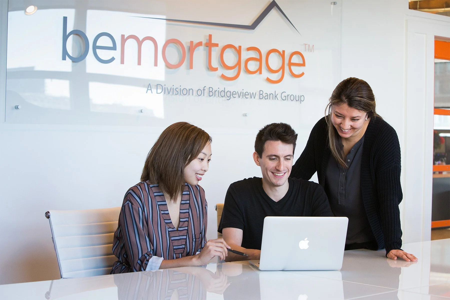Three people gathered around a laptop, smiling and working together in an office with a sign that reads 'bemtorage, a division of Bridgeview Bank Group.'