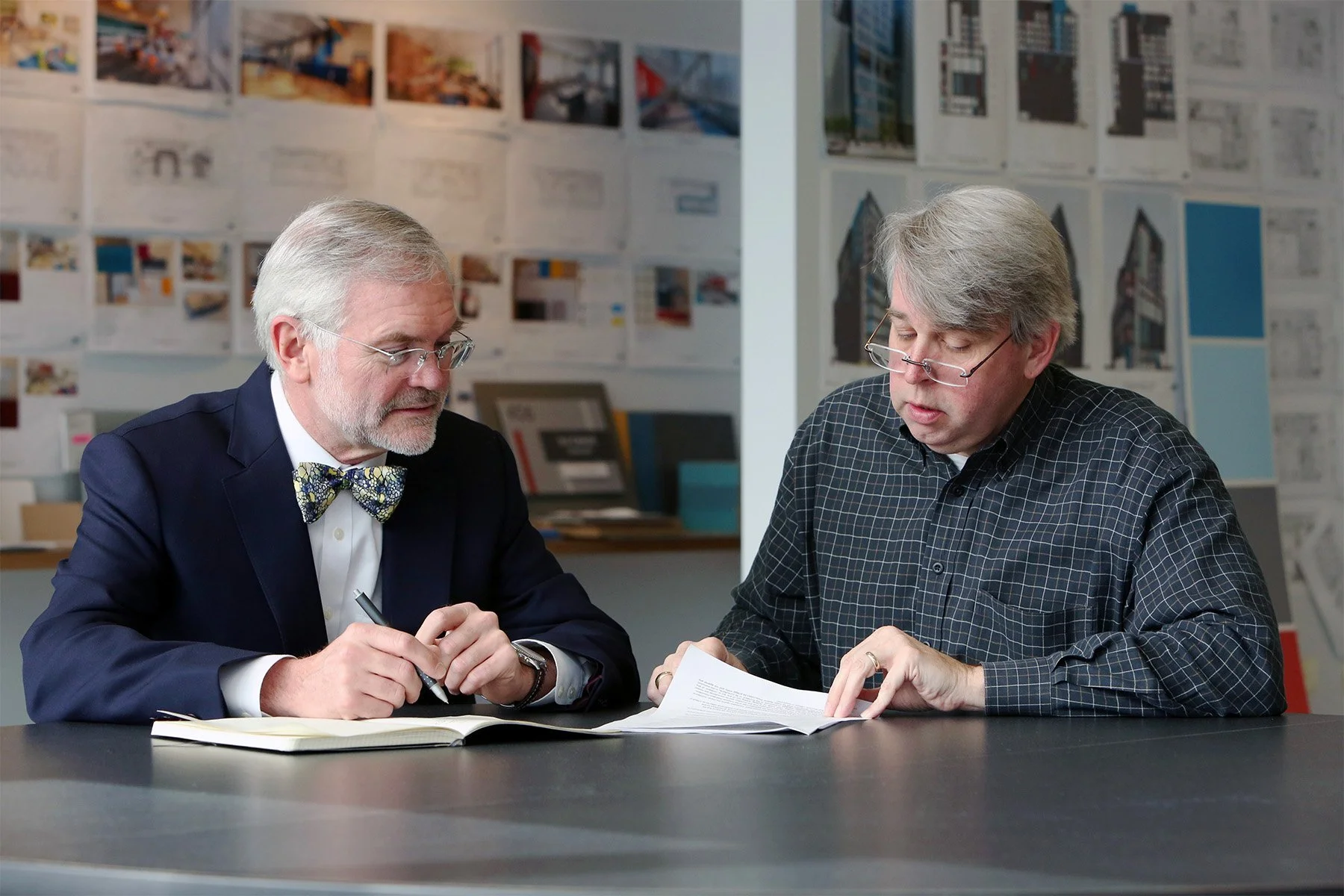 Two middle-aged men with gray hair and glasses sitting at a table in an office, reviewing documents together. One man is wearing a dark suit and bow tie, the other in a checkered shirt. The background has walls decorated with architectural or design 