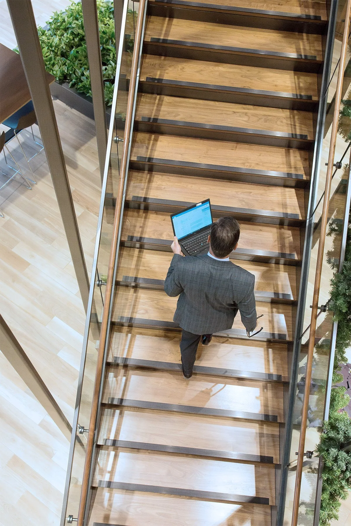 A man in a suit walking up a wooden staircase while holding a laptop and glasses in his right hand.
