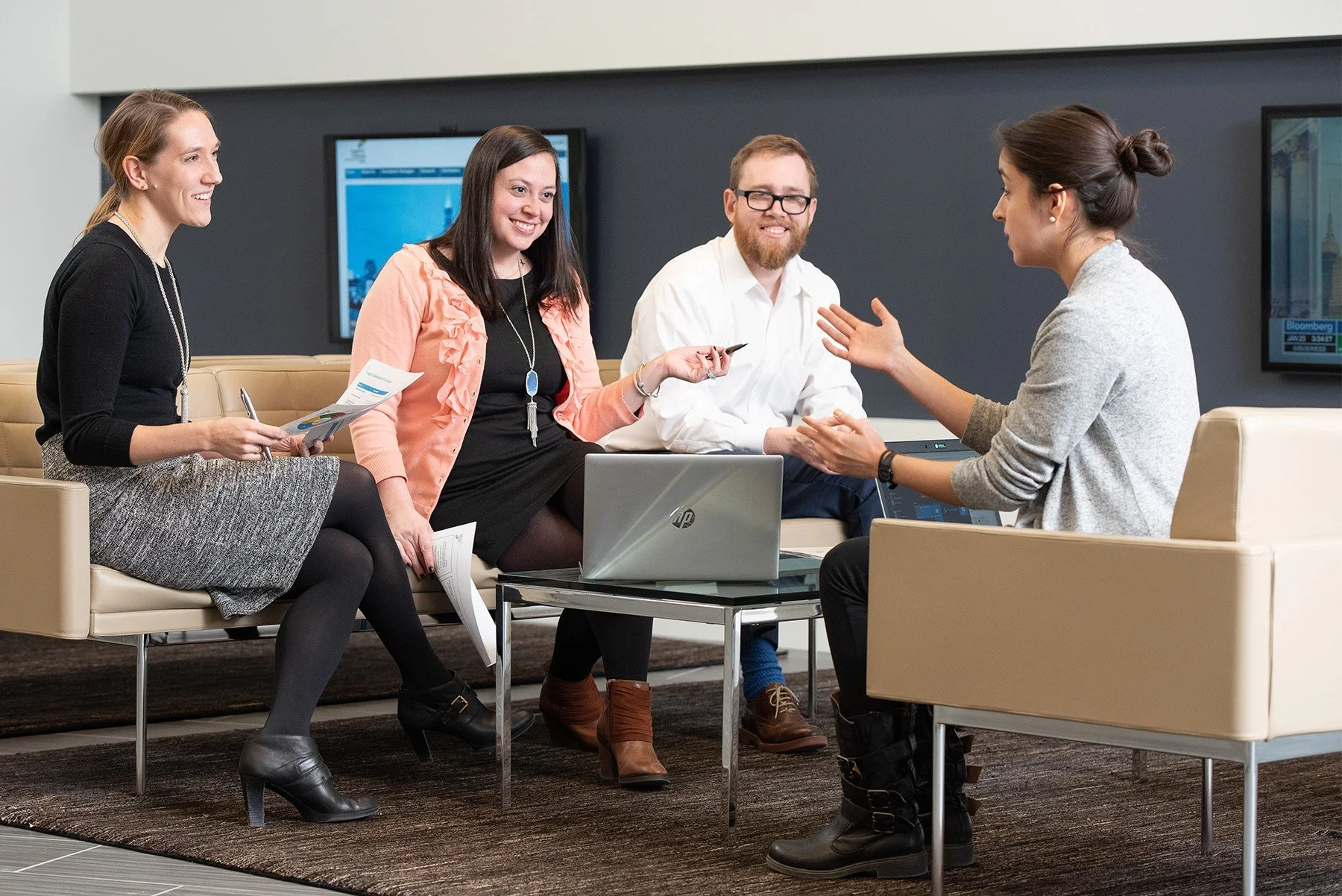 Four people sitting on a beige sofa in a modern office, engaged in a discussion with one woman speaking and three listening, with laptops and papers on their laps.