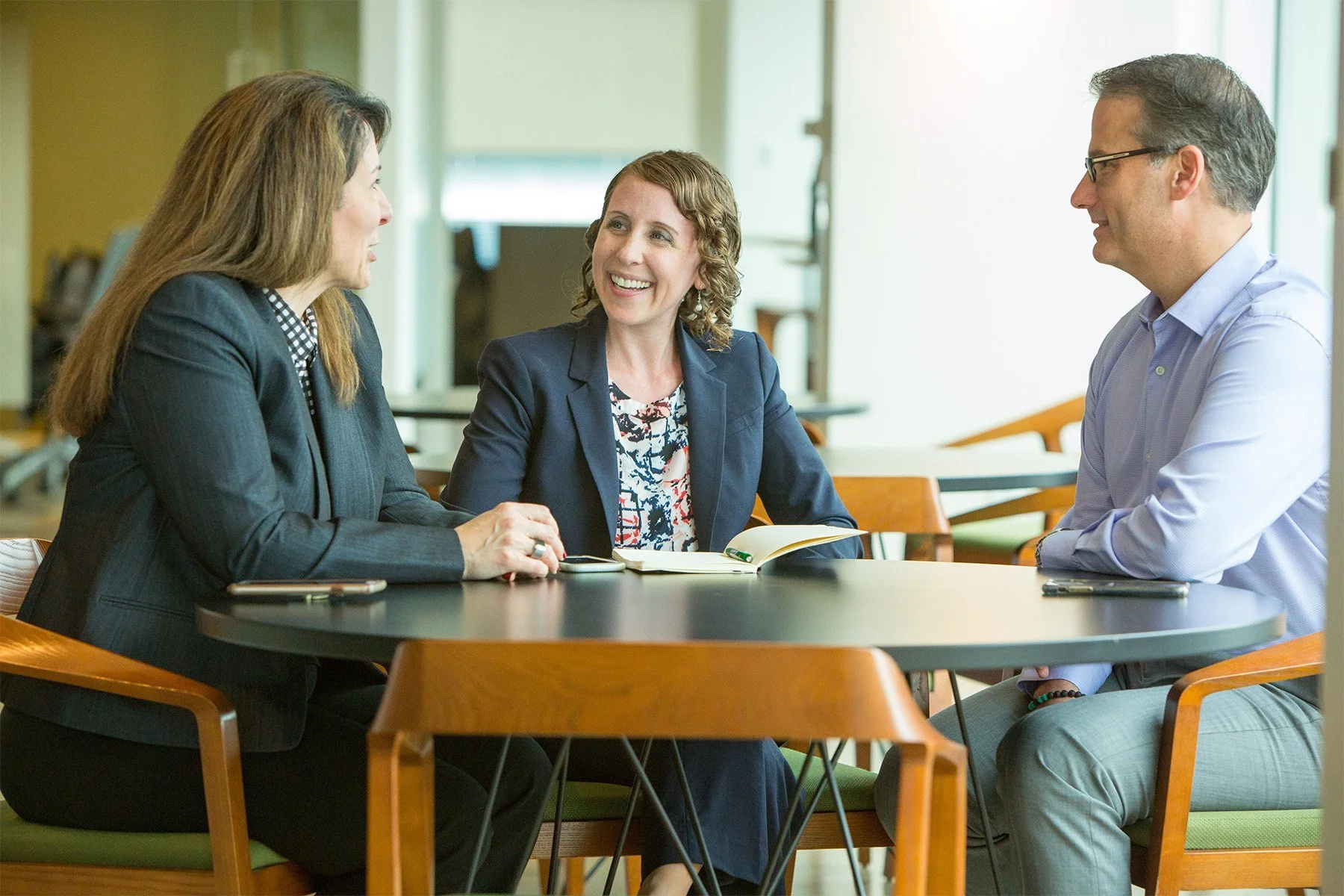 Three professionals, two women and one man, sitting at a round table in a well-lit office, engaged in conversation and smiling.