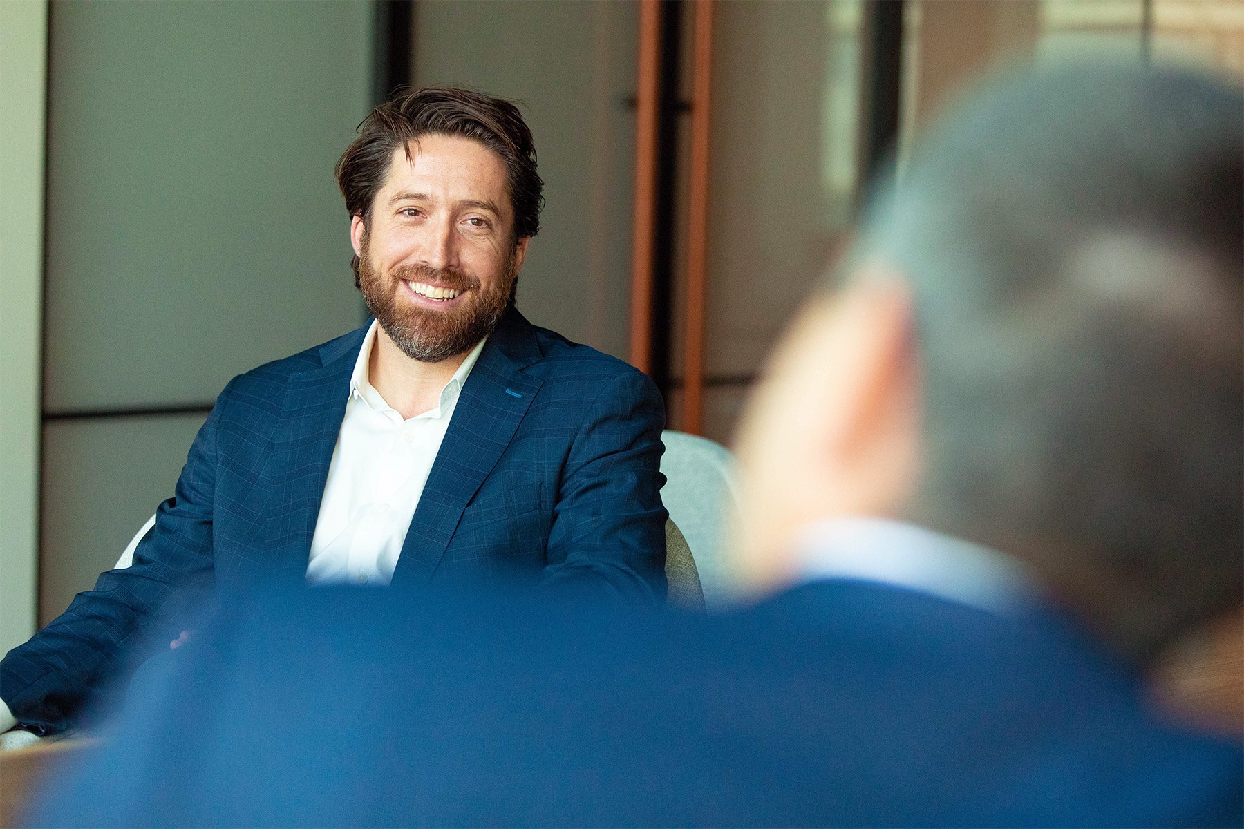 A man in a blue plaid suit with a white shirt, smiling and sitting at a table during a conversation, with another person visible in the foreground.