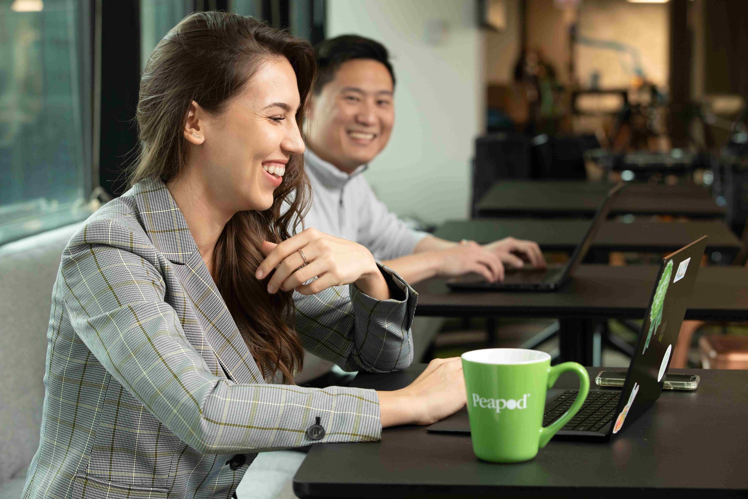 A smiling woman with long brown hair wearing a plaid blazer is sitting at a black table in a meeting room, working on a laptop. A man with short black hair, wearing a white shirt, is sitting next to her, also smiling and working on his laptop. There 