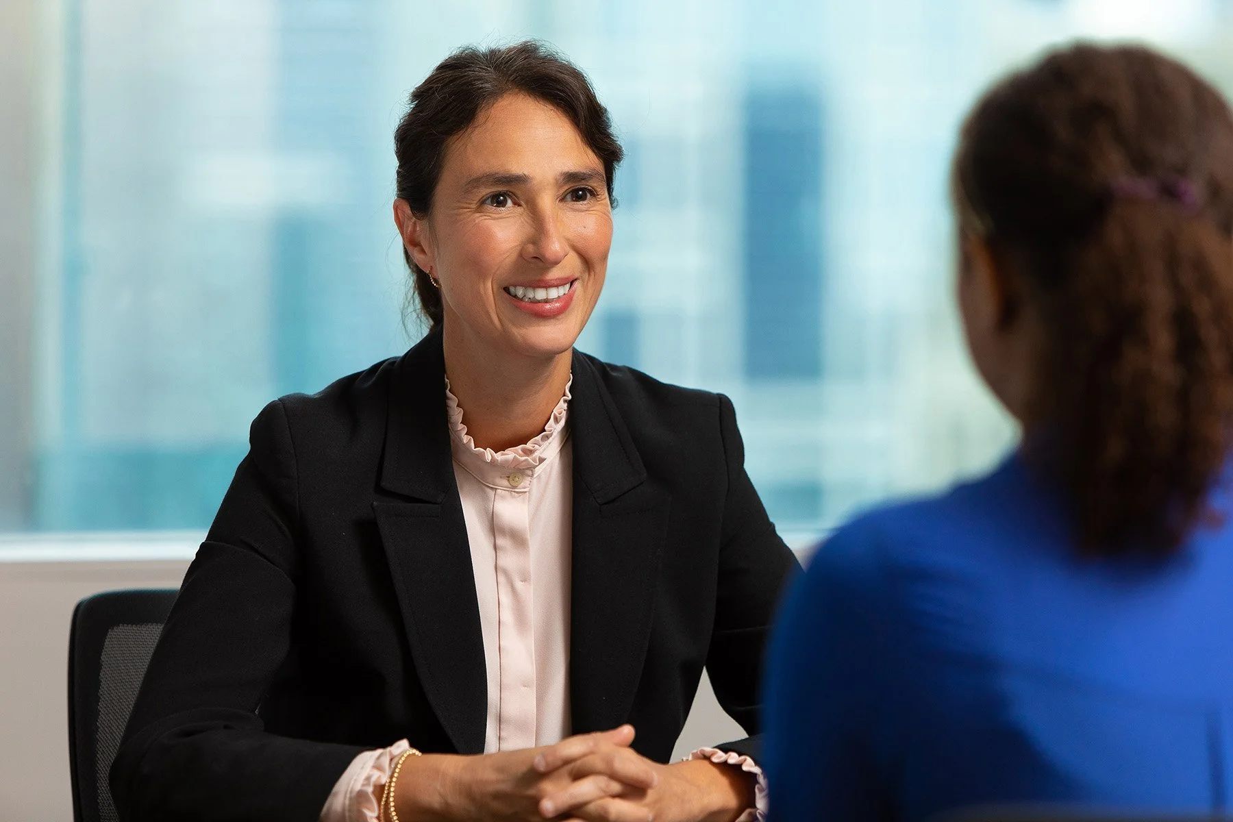A woman with short dark hair, wearing a black blazer and light-colored blouse, smiling and talking to another woman with curly hair, wearing a blue top, in a professional setting with a glass window in the background.
