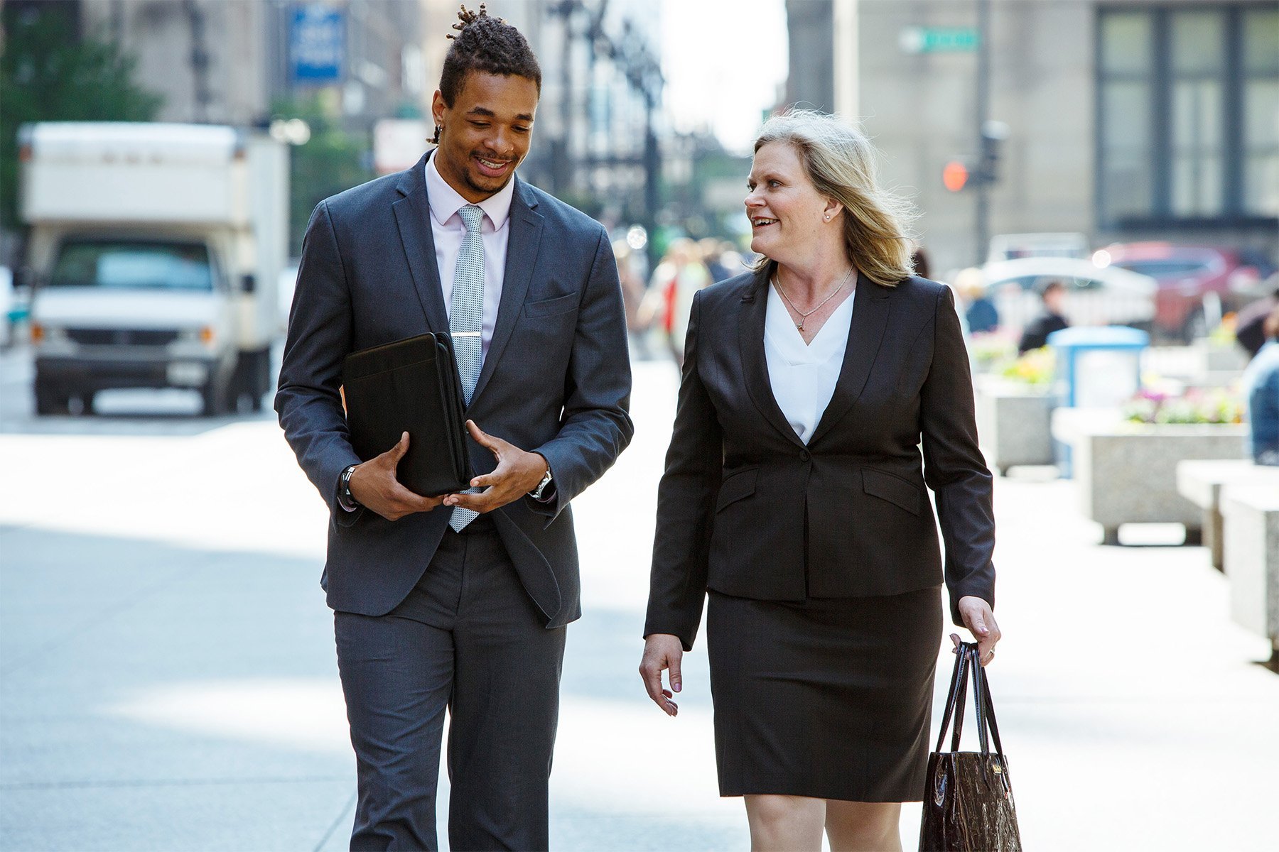 Two professionals, a man and a woman, walking and talking outdoors in an urban setting. The man is smiling, wearing a suit and holding a folder. The woman is wearing a business suit, smiling, and holding a shopping bag.