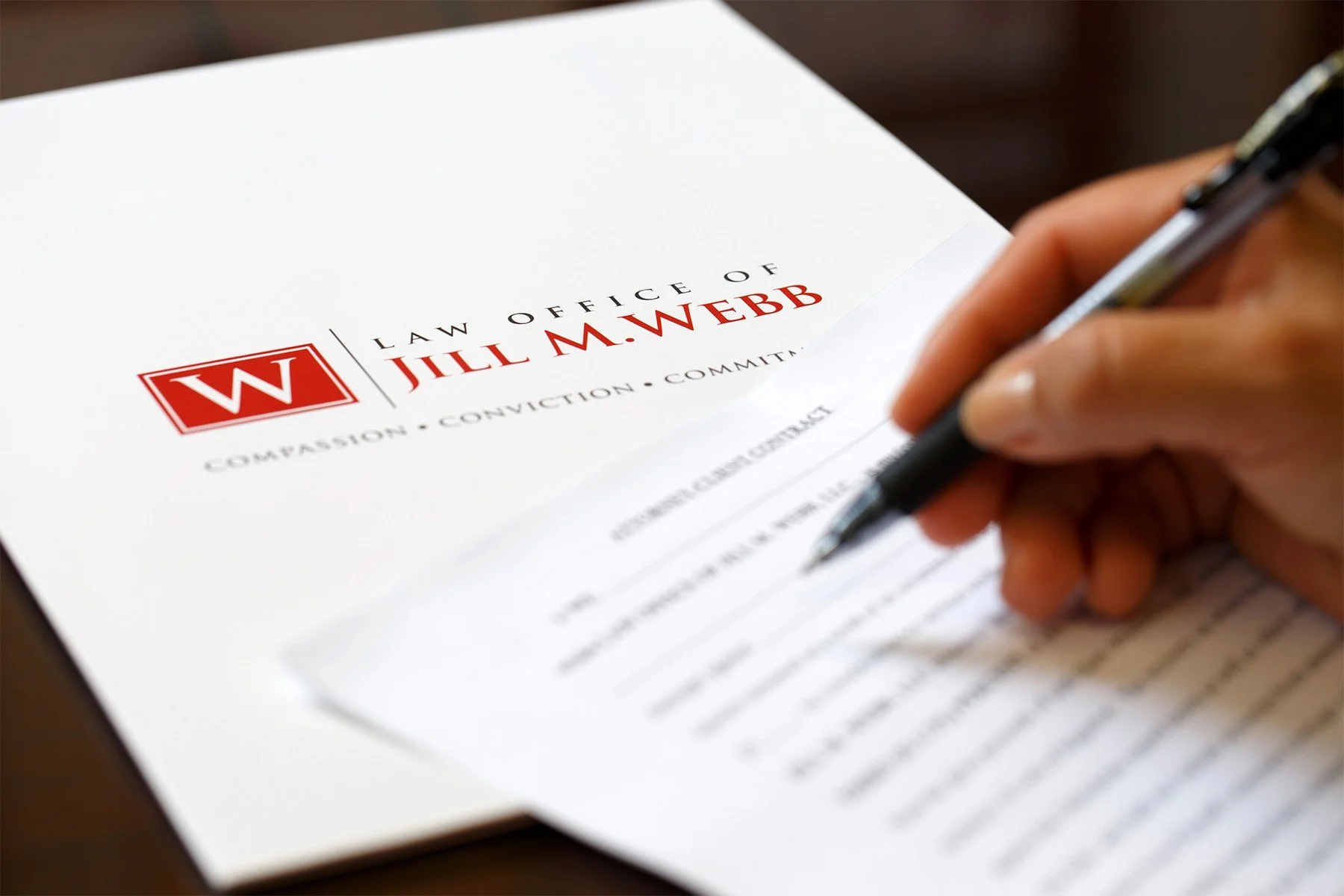 A person holding a pen and signing a legal document from the Law Office of Jill M. Webb.