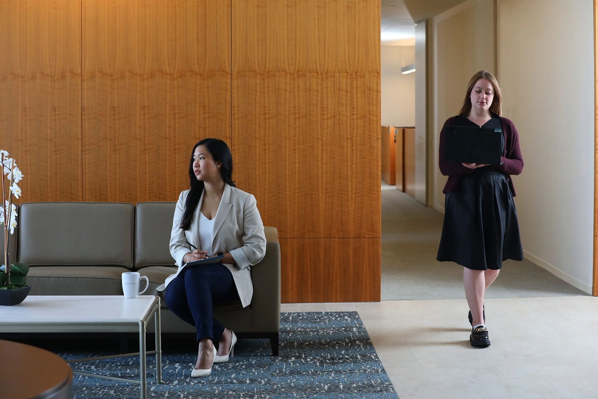 Two women in a professional setting. One woman with long black hair, wearing a light-colored blazer and dark pants, is sitting on a beige sofa taking notes. The other woman with shoulder-length light brown hair, wearing a dark skirt and a maroon card