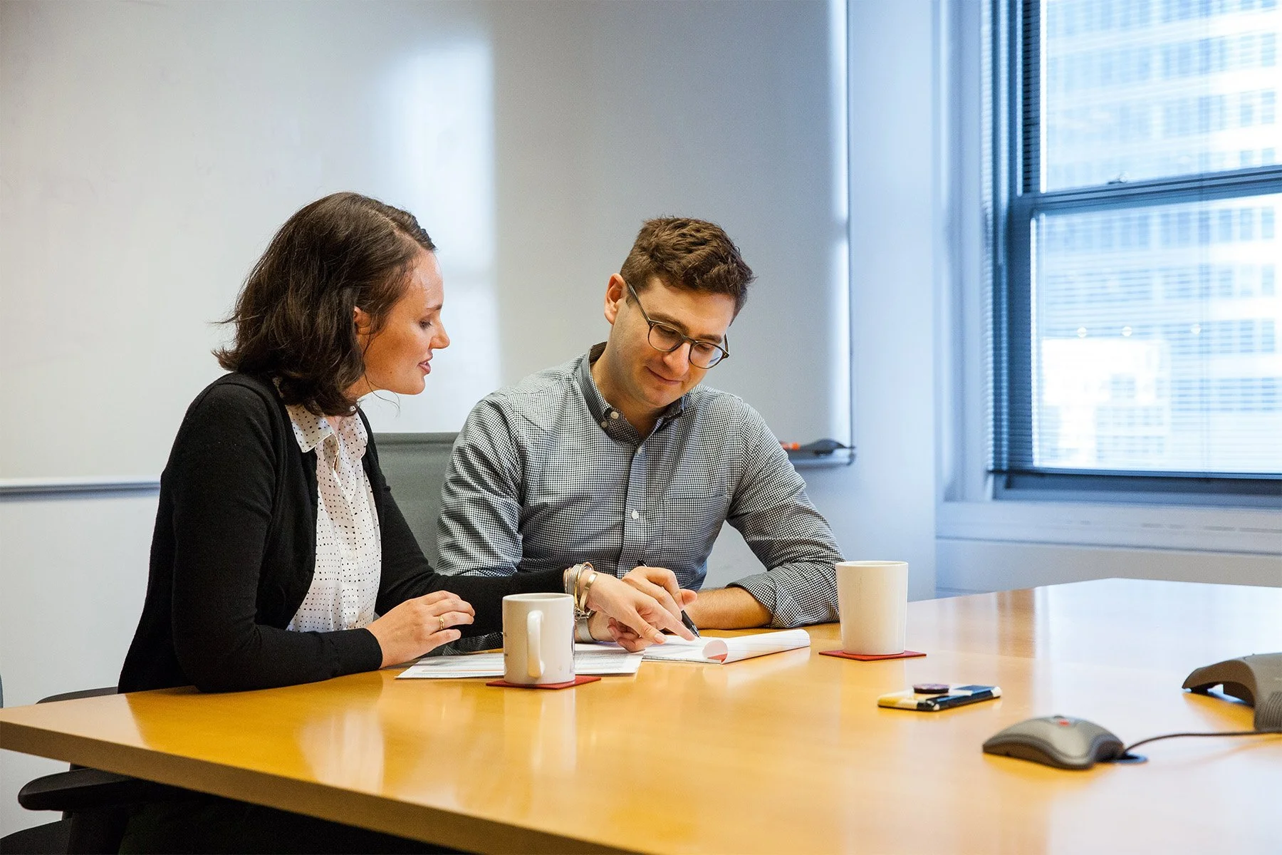 Two colleagues, a woman and a man, sitting at a conference table in a modern office, reviewing documents and discussing work.