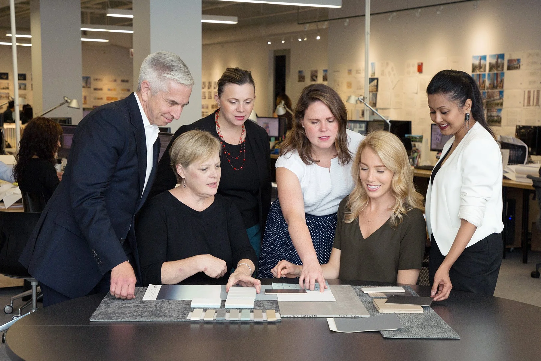 A group of six women and one man gathered around a table examining samples and color swatches, with a bright open office setting in the background.