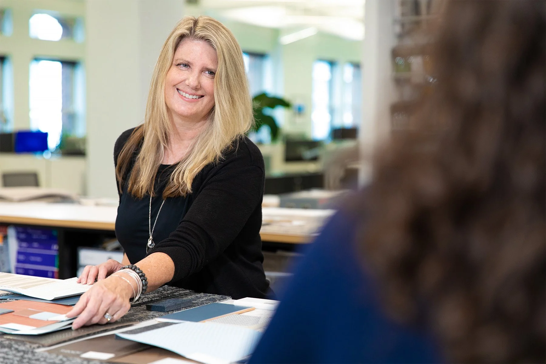 A woman with long blonde hair smiling while sitting at a table with color samples and design materials, engaging in a discussion in a bright office space.