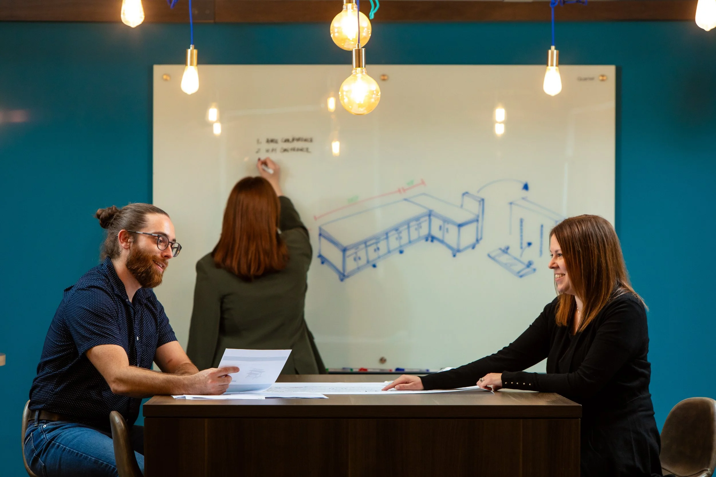 Two women and one man in a business meeting, with a woman writing on a whiteboard in the background. The setting appears to be a modern conference room with hanging lights and a teal wall.