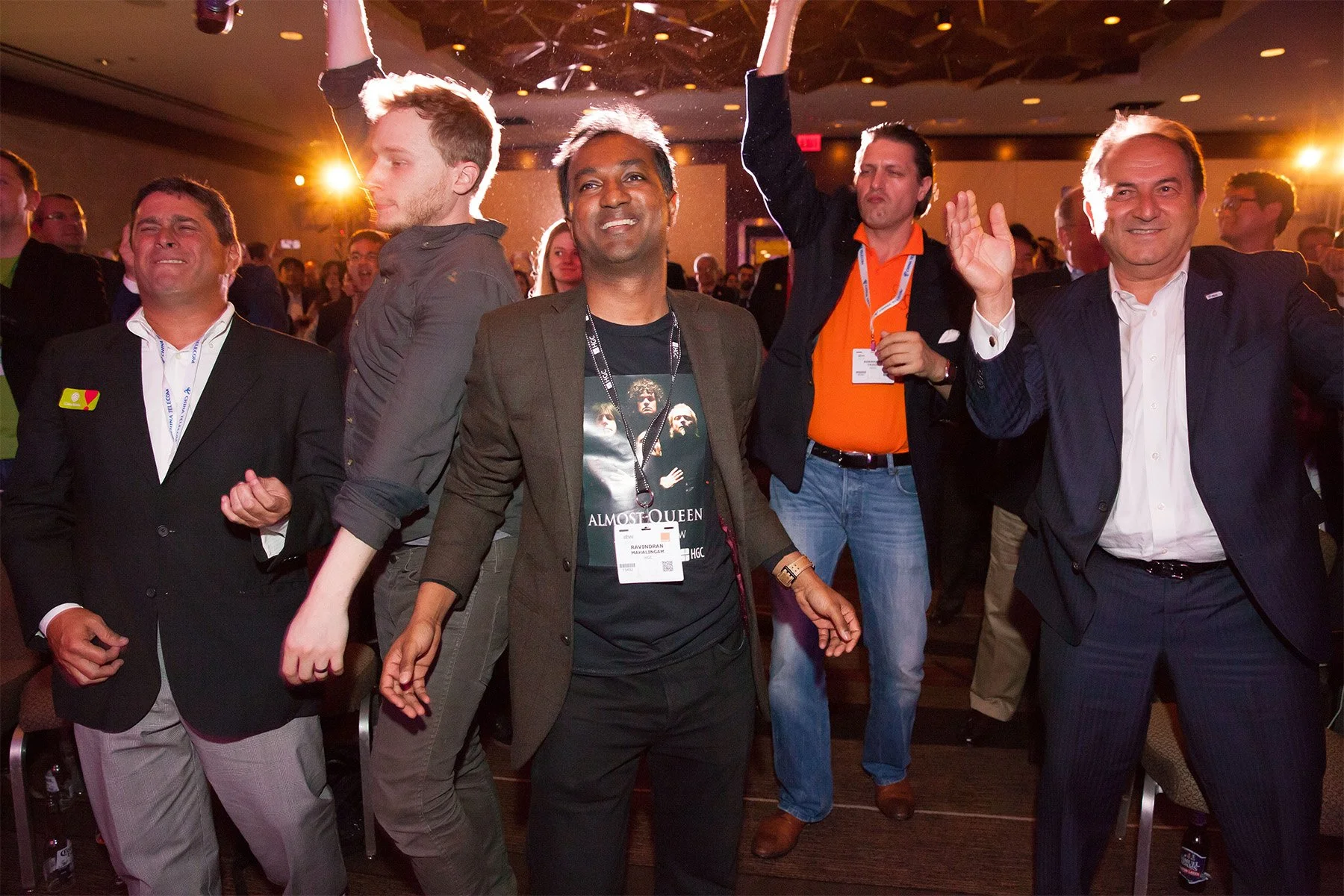 A group of five men at a conference or event, with some holding hands or raising their arms, in a crowded room with warm lighting.