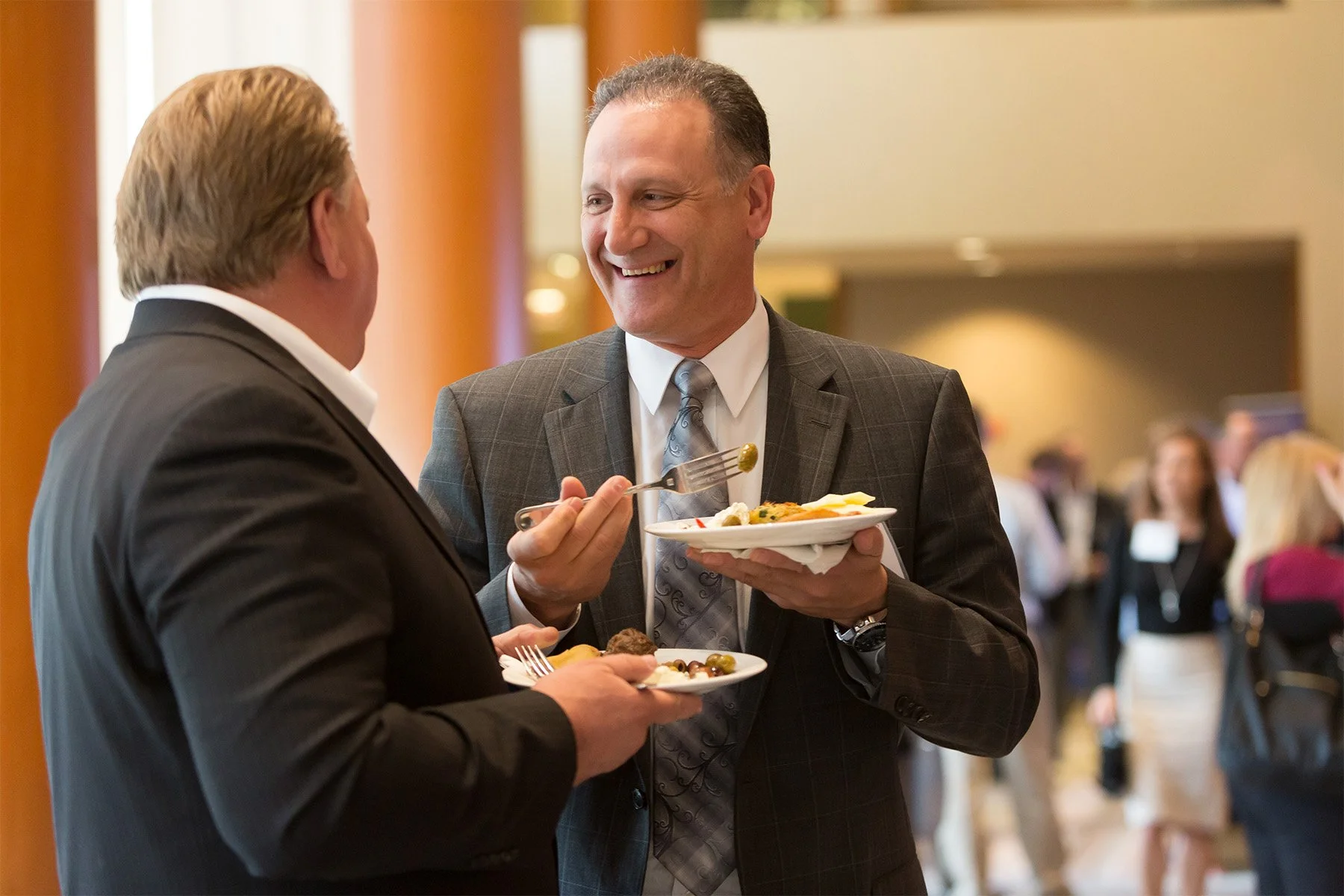 Two men in business suits smiling and talking at a social event, each holding a plate of food. The background shows other attendees and a warm, well-lit setting.