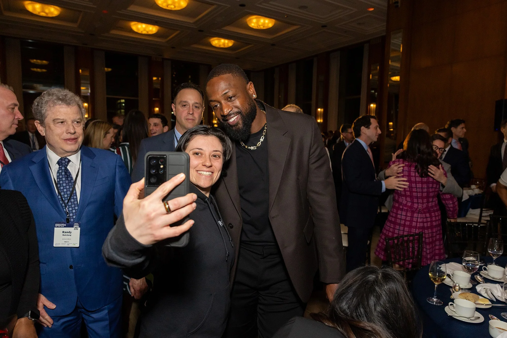 People gathered at a formal event, including a woman taking a selfie with a man. The woman has short dark hair and is smiling, while the man has a beard, is dressed in a black suit with a chain necklace, and is also smiling. Surrounding them are othe