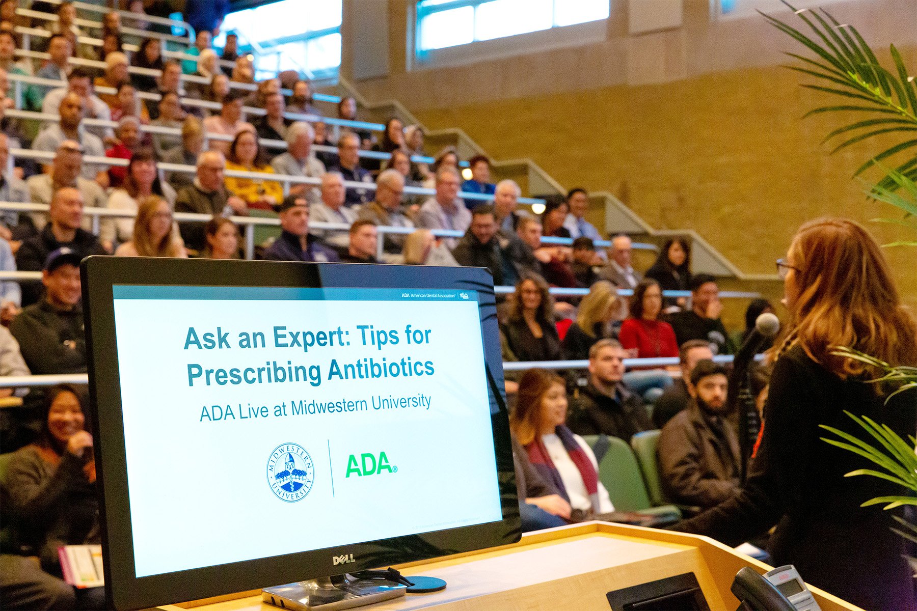Lecture hall filled with audience listening to a speaker. A computer screen in the foreground reads, 'Ask an Expert: Tips for Prescribing Antibiotics,' with logos for Midwest University and ADA.