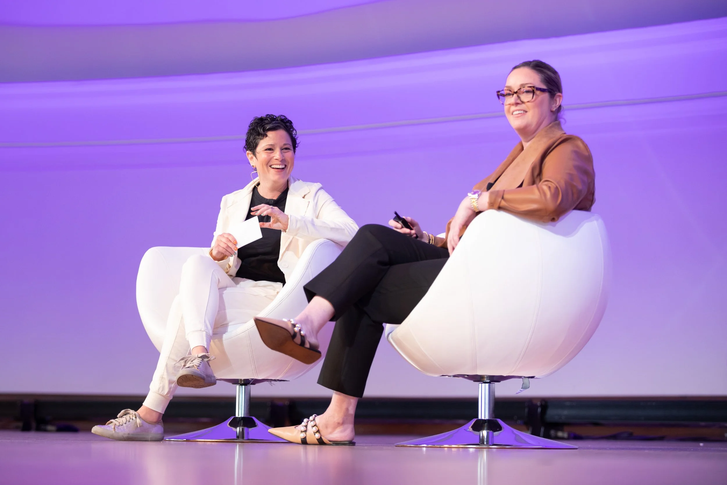 Two women sitting on stage in white chairs, smiling and engaging in conversation, with a purple background.