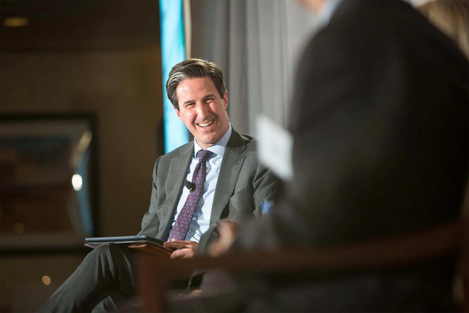 A man in a gray suit and patterned tie smiling and holding a tablet during a discussion or interview, with someone else partially visible in the foreground.