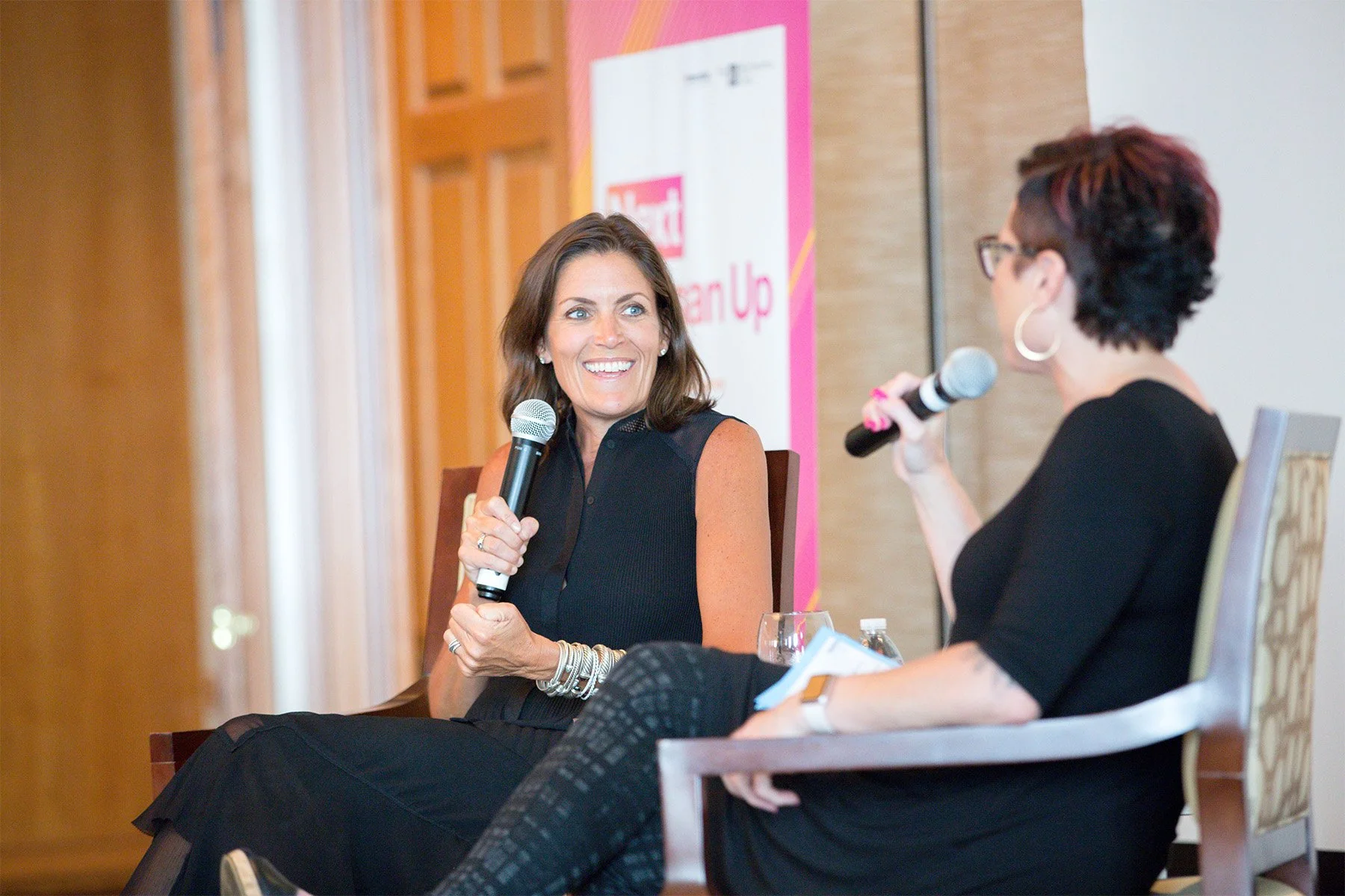 Two women speaking into microphones during a panel discussion at an event. One woman, with brown hair, is smiling and wearing a sleeveless black dress, while the other woman has short dark hair, glasses, and is holding notes.