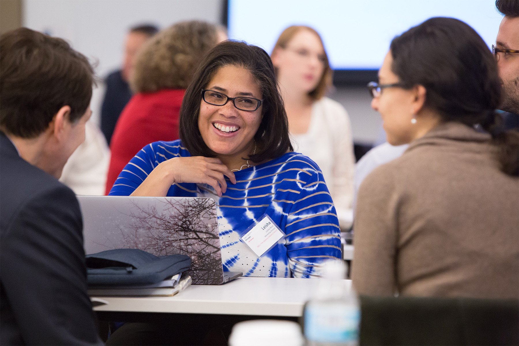 Group of diverse professionals engaging in a discussion at a conference table, with a woman smiling and wearing glasses in focus.