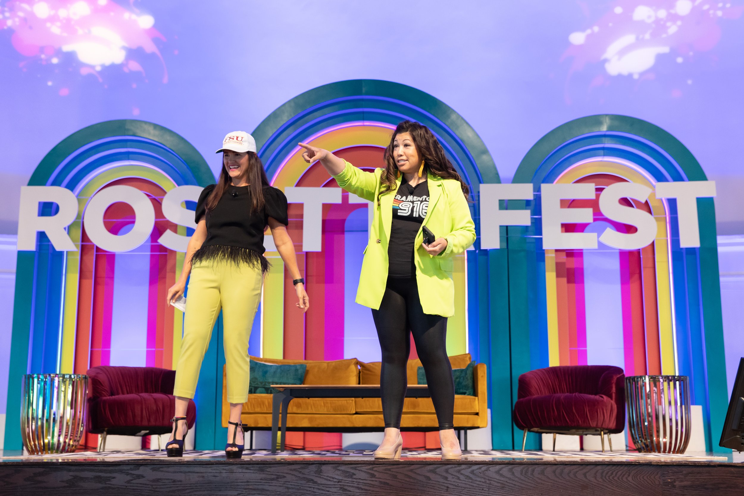 Two women on a colorful stage with large letters spelling 'ROSTFEST' and rainbow arches background. One woman wears a yellow pants and black top, the other a neon green blazer and black pants, both smiling and engaged in conversation.