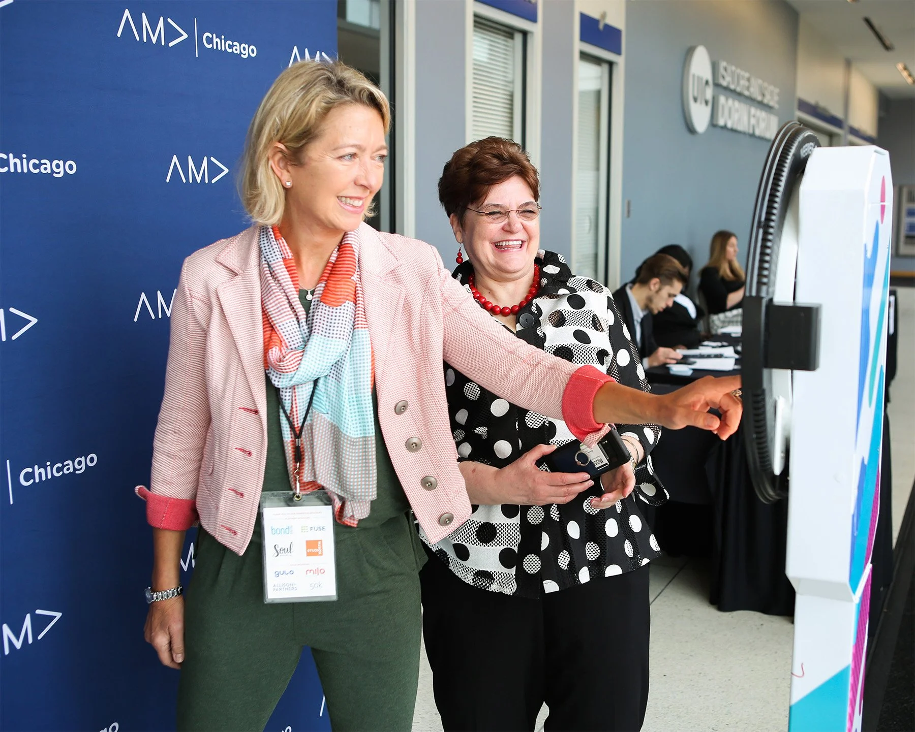 Two women smiling and taking a selfie at a conference or event with a dark blue backdrop that reads 'AM' and 'Chicago'. Other attendees are visible in the background.