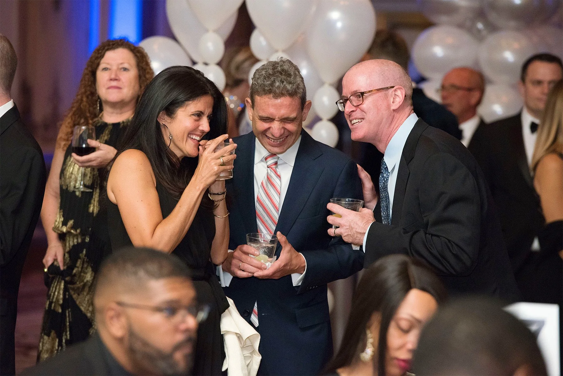 A group of people at a formal event, some holding drinks, smiling and laughing together. In the background, there are white balloons and other guests mingling.
