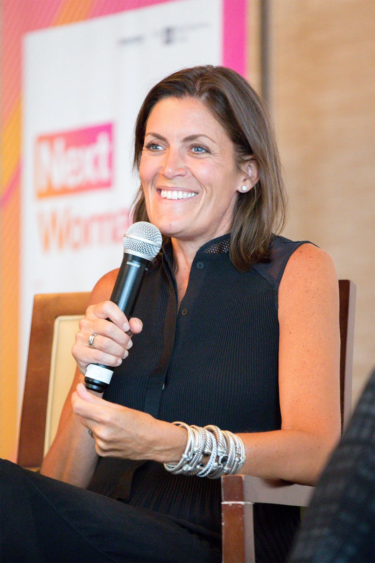 A woman with brown hair and blue eyes smiling and holding a microphone, sitting on a chair during a discussion or interview.