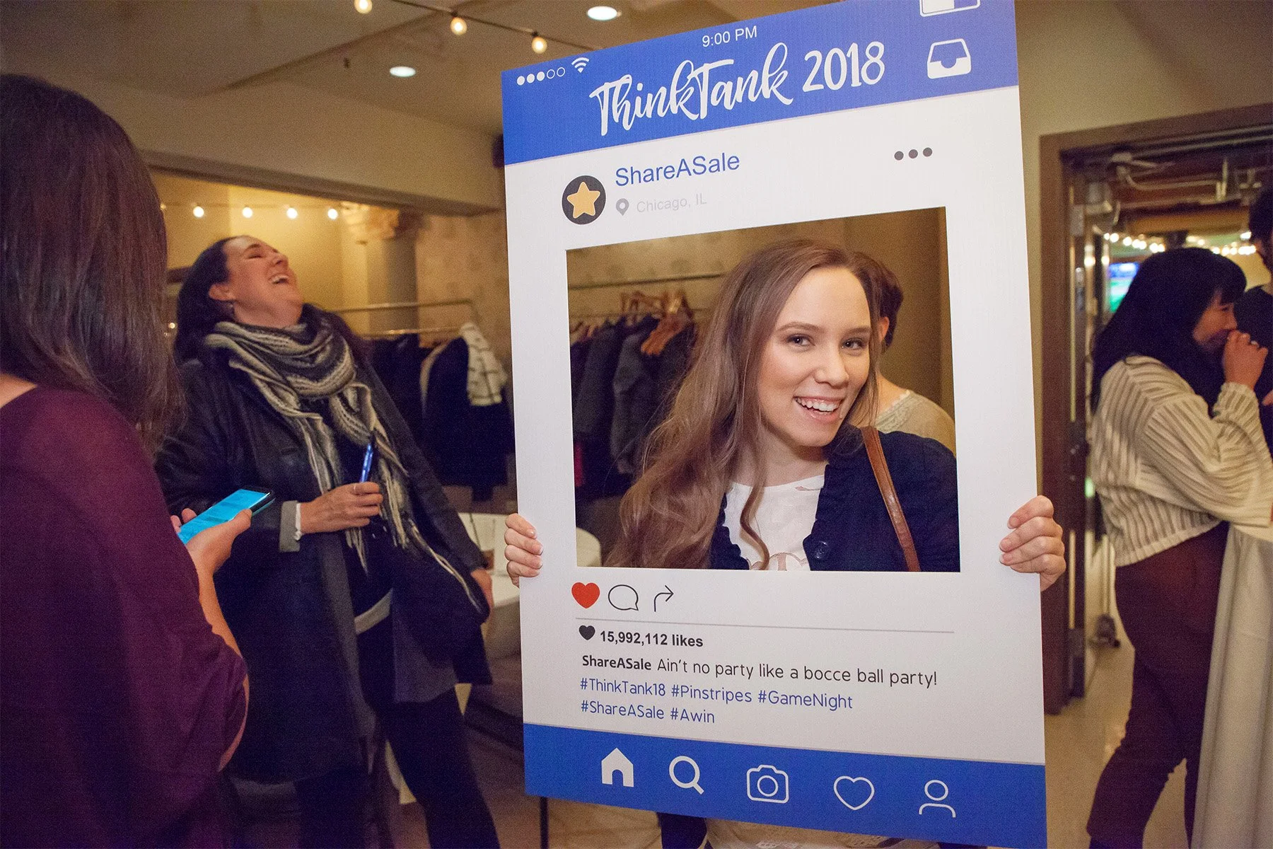 A woman holding a large Instagram photo frame prop at an event, with other women in the background. The frame displays a social media post from ThinkTank 2018 with a picture of a smiling young woman. The event is held indoors with string lights overh