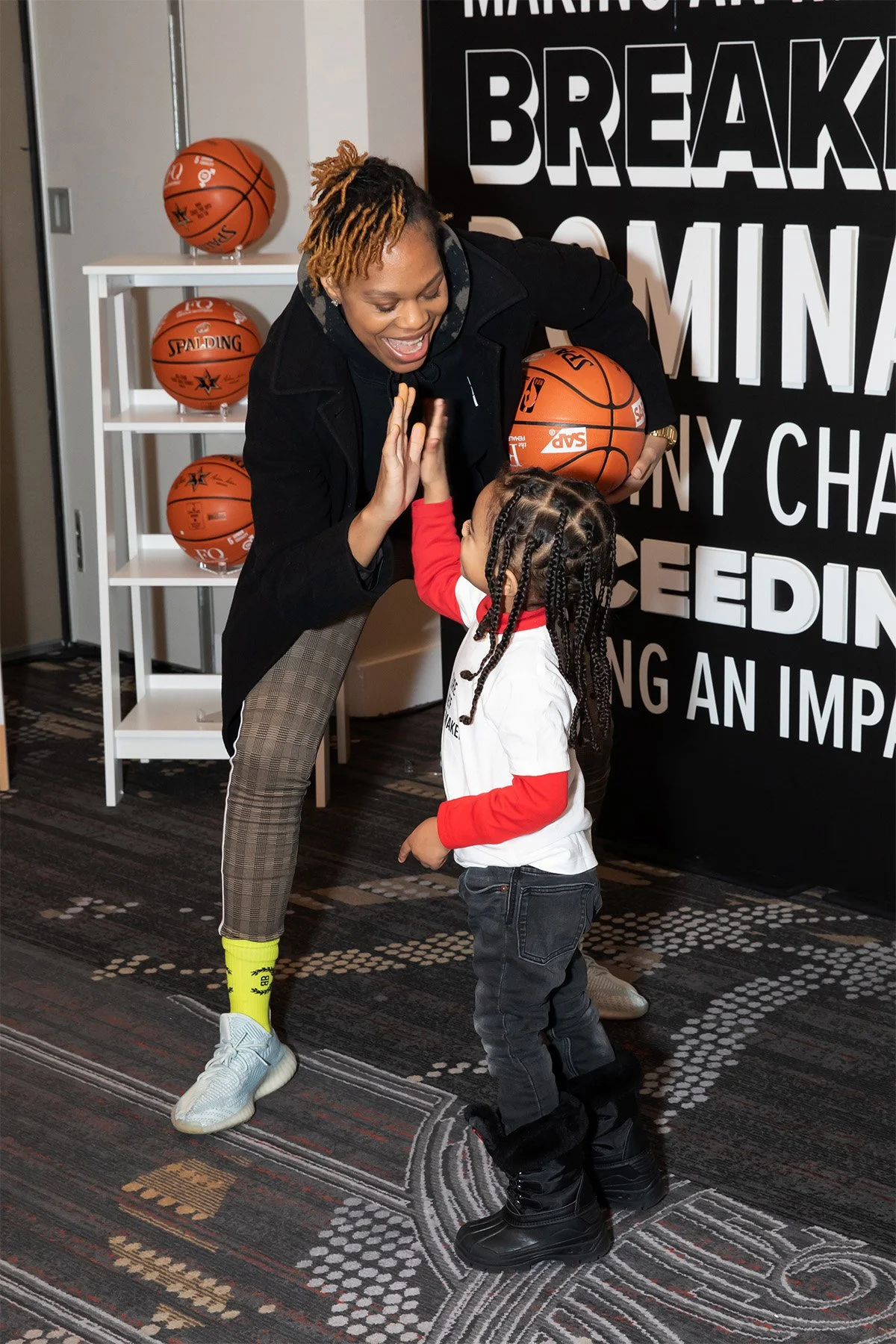 A woman and a young girl high-fiving in a room with basketballs and a large black poster with white lettering in the background.