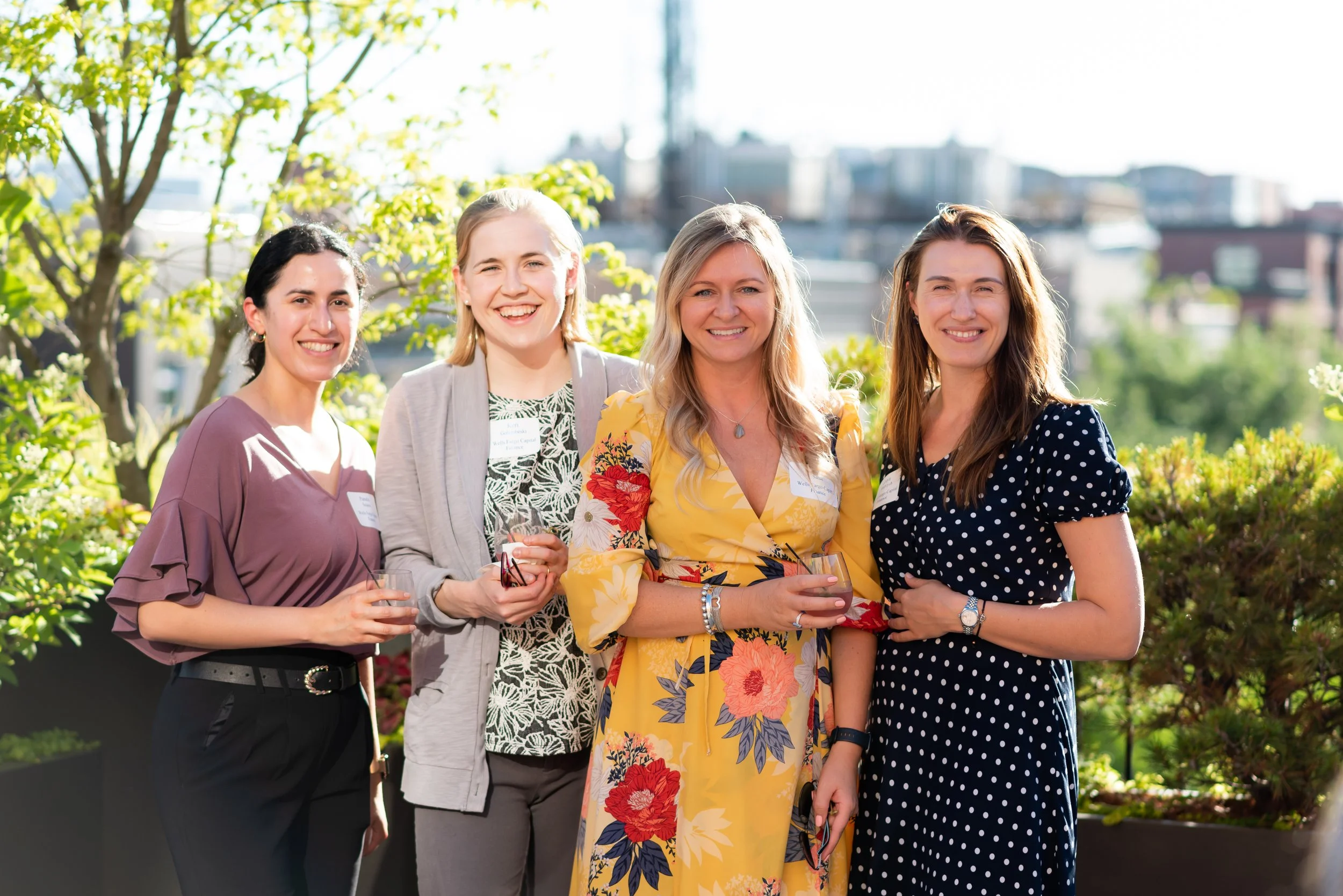 Group of five women smiling and holding drinks at an outdoor gathering on a sunny day, with city buildings and green trees in the background.