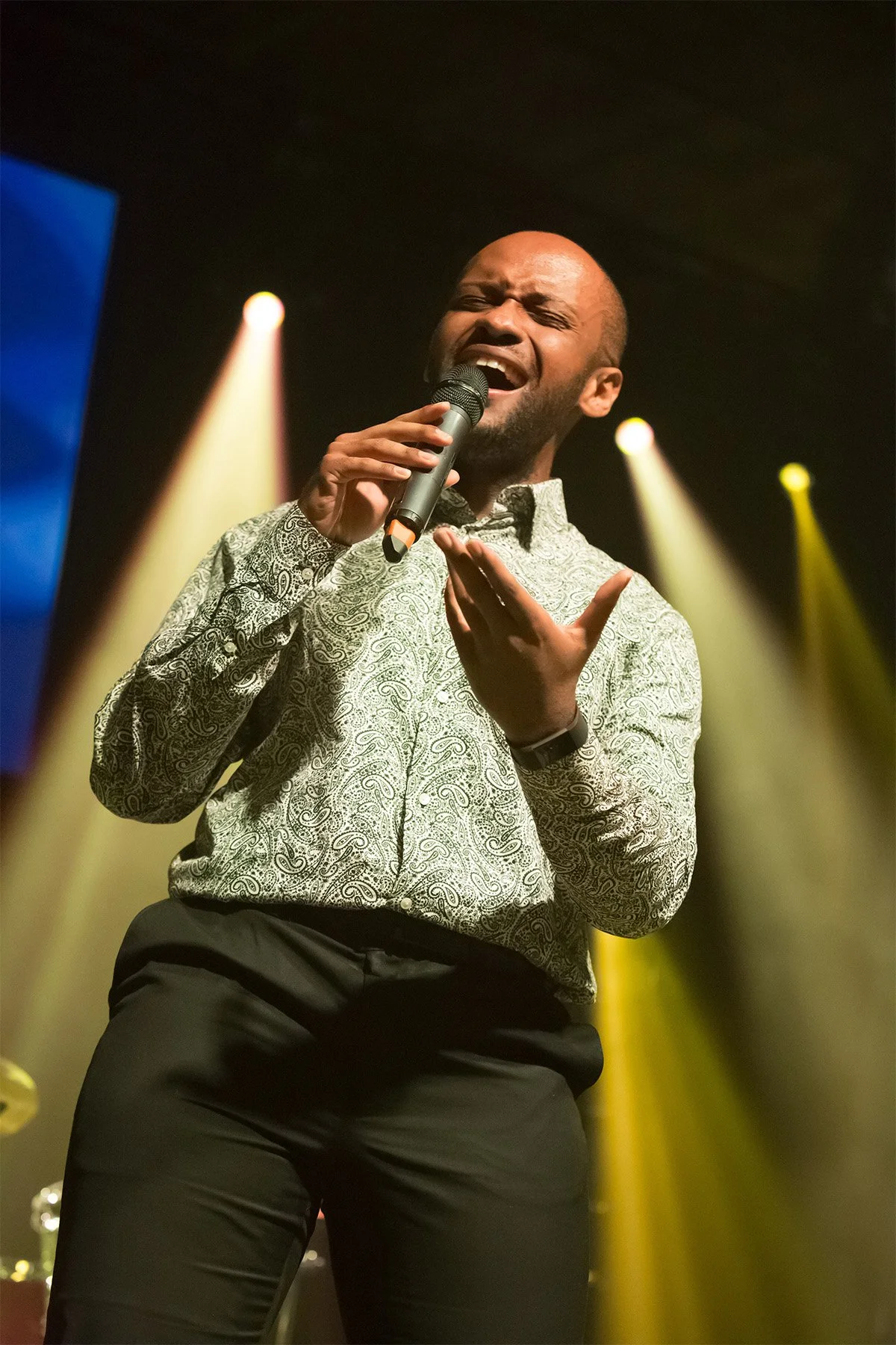 A man singing passionately on stage, holding a microphone with one hand and gesturing with the other, illuminated by colorful stage lights.
