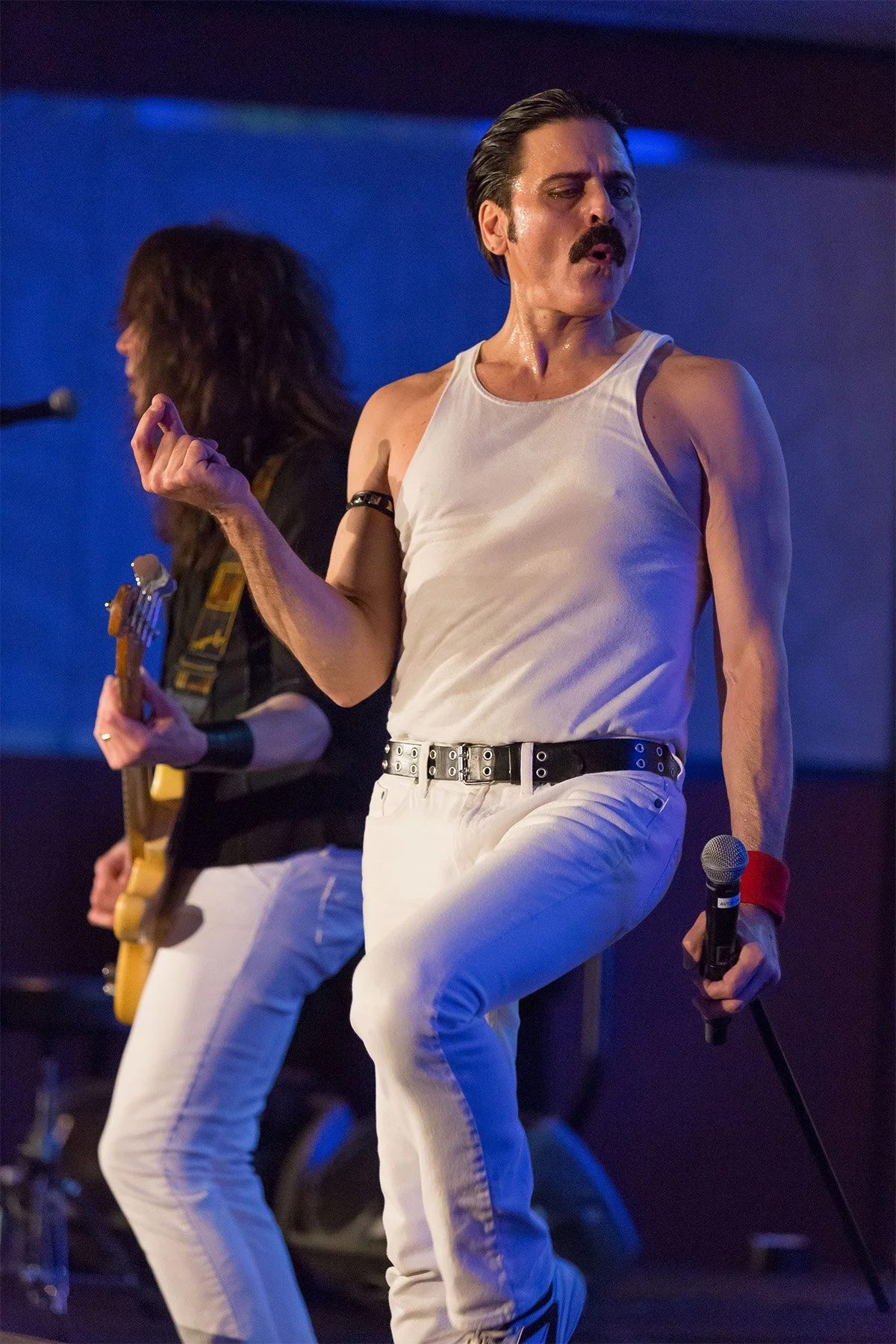 Musical performers on stage, one singing into a microphone and the other playing guitar, both wearing white jeans and tank tops.