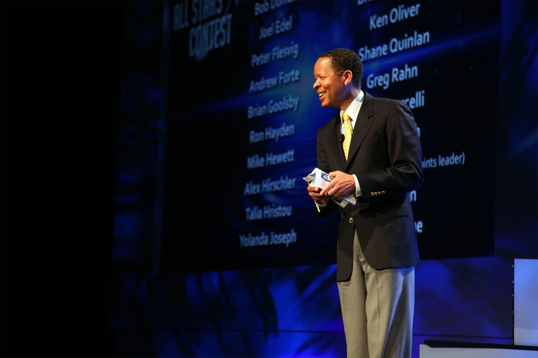 A man in a dark blazer and light-colored pants stands on a stage, smiling and holding papers. Behind him is a large screen displaying a list of names and the title 'Best Games'. The background is dark with blue lighting.