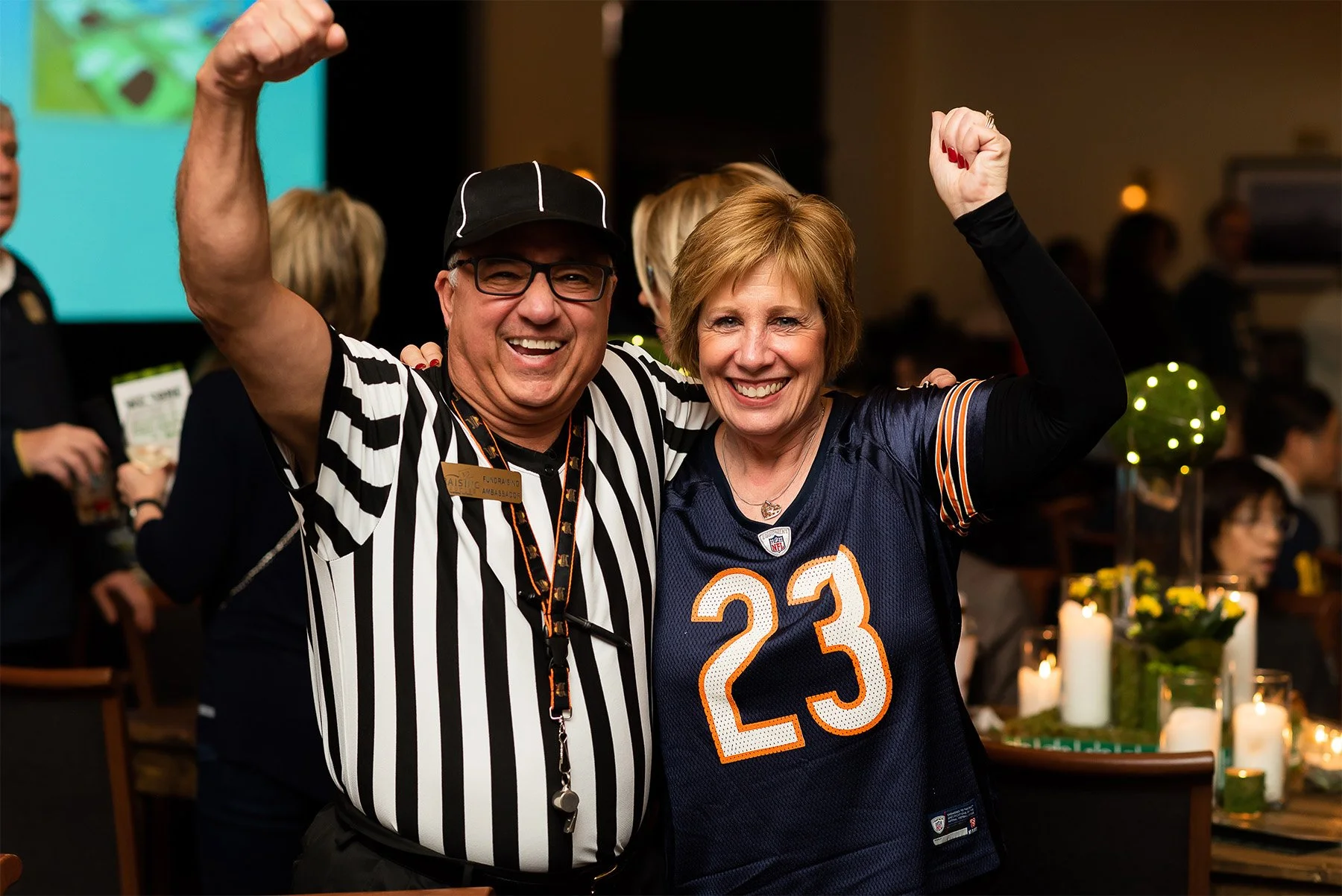 A man dressed as a sports referee and a woman wearing a Chicago Bears football jersey, both smiling and raising their arms in celebration at an indoor event with candles and decorations in the background.