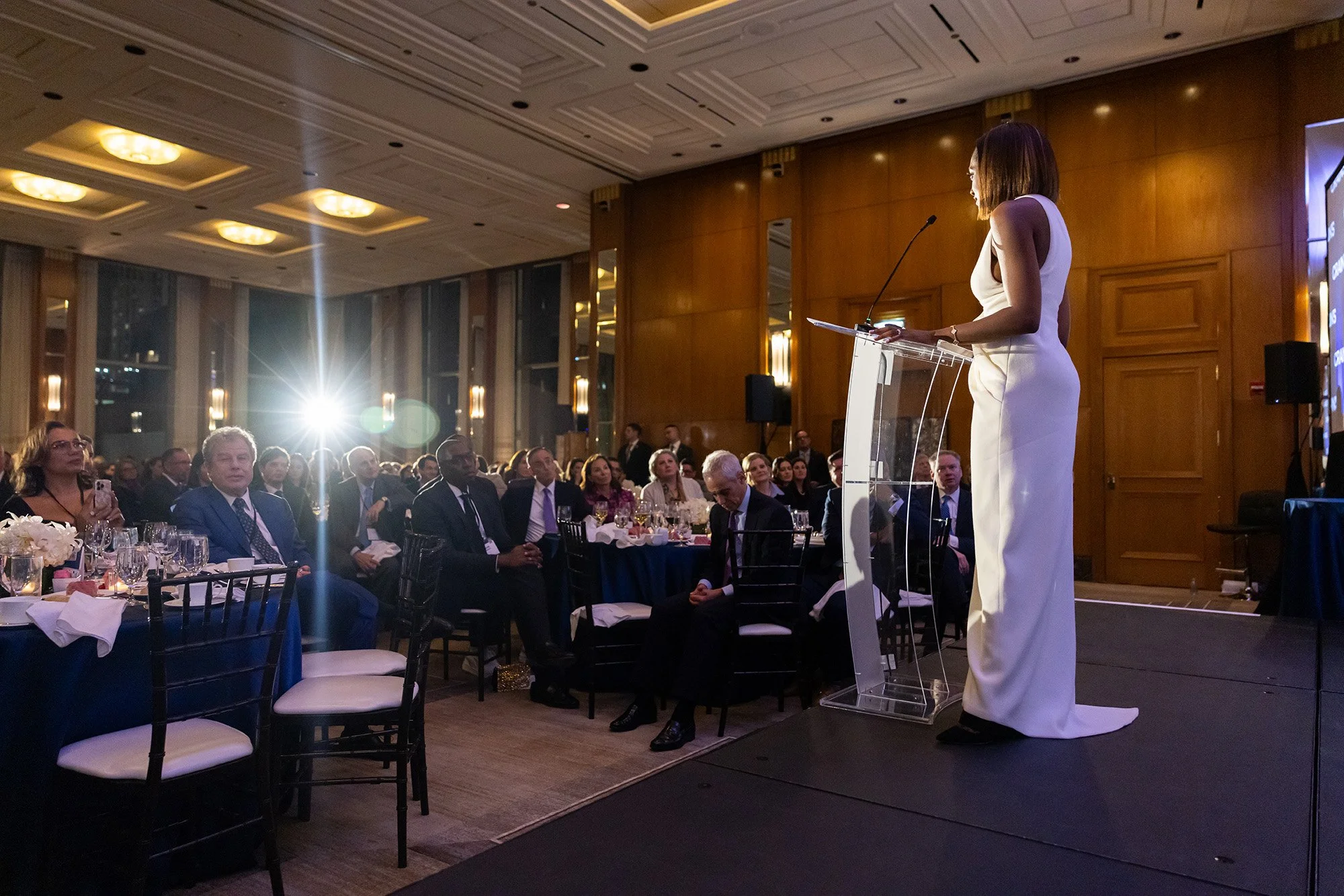 A woman in a white dress giving a speech at a formal event or conference, standing behind a clear podium, with a seated audience in front of her. The room has warm lighting and wood-paneled walls.