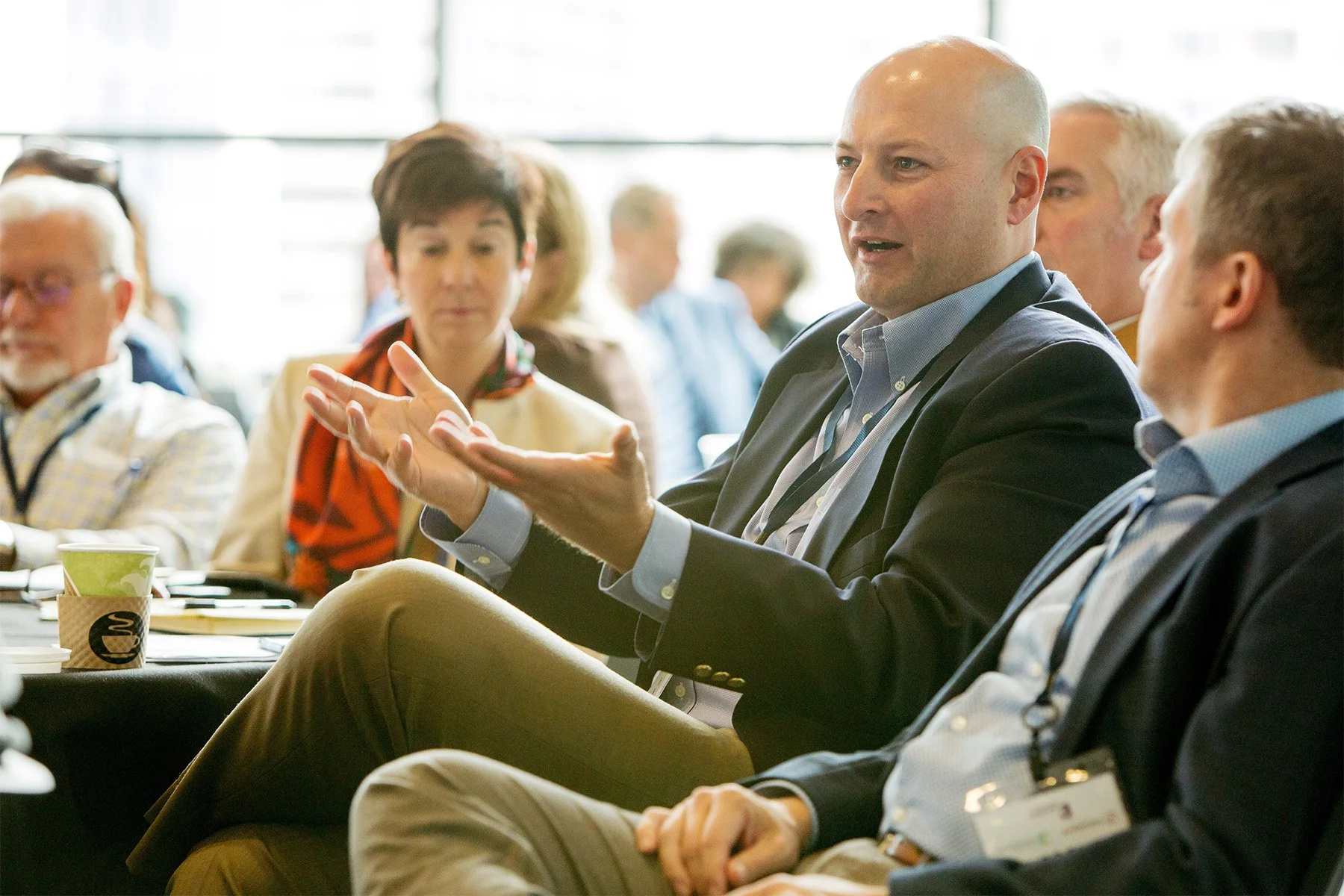 Man in a blazer speaking and gesturing during a panel or discussion at a conference with other attentive participants.