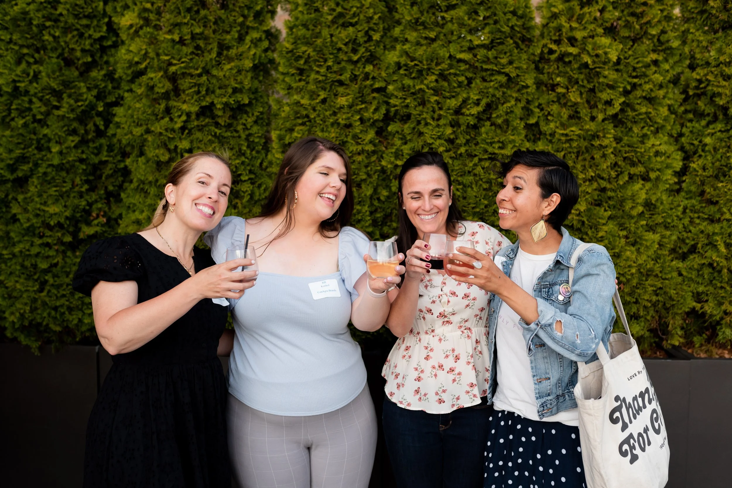 Four women smiling and holding glasses of wine while standing together outdoors in front of a green hedge.