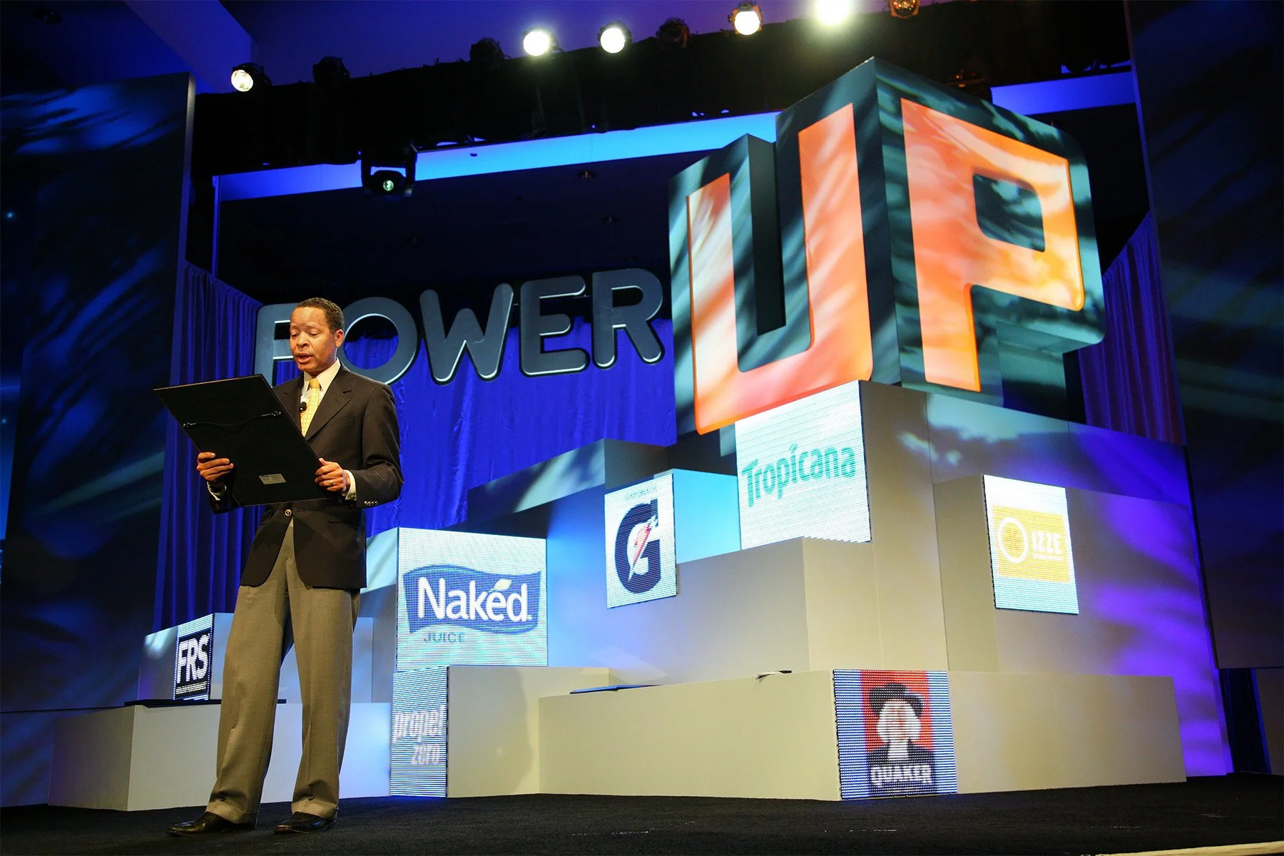 A man in a suit and tie stands on stage reading from a black folder at a conference or presentation. Behind him is a large colorful display with the word 'POWER' and the letters 'UP' in large 3D format. The stage is decorated with multiple logos and 