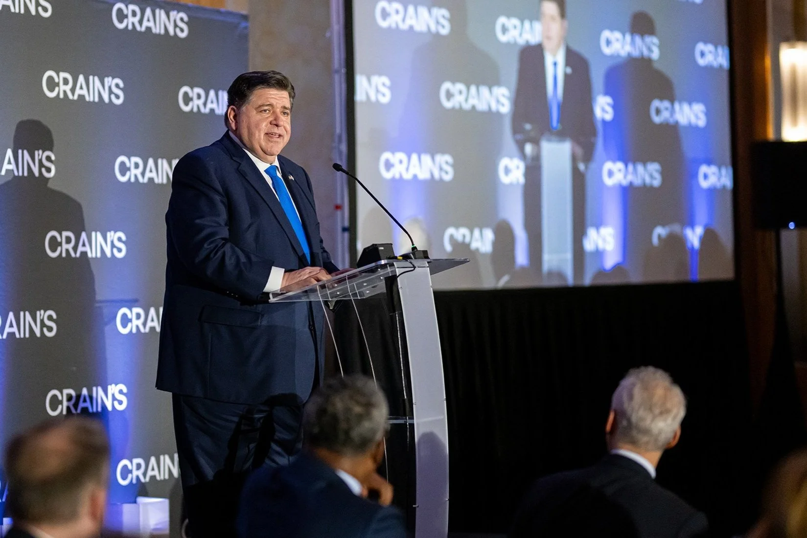 A man in a dark suit and blue tie is speaking at a podium during a conference, with a large screen behind him displaying his image and the word 'CRAIN'S' repeatedly in the background.