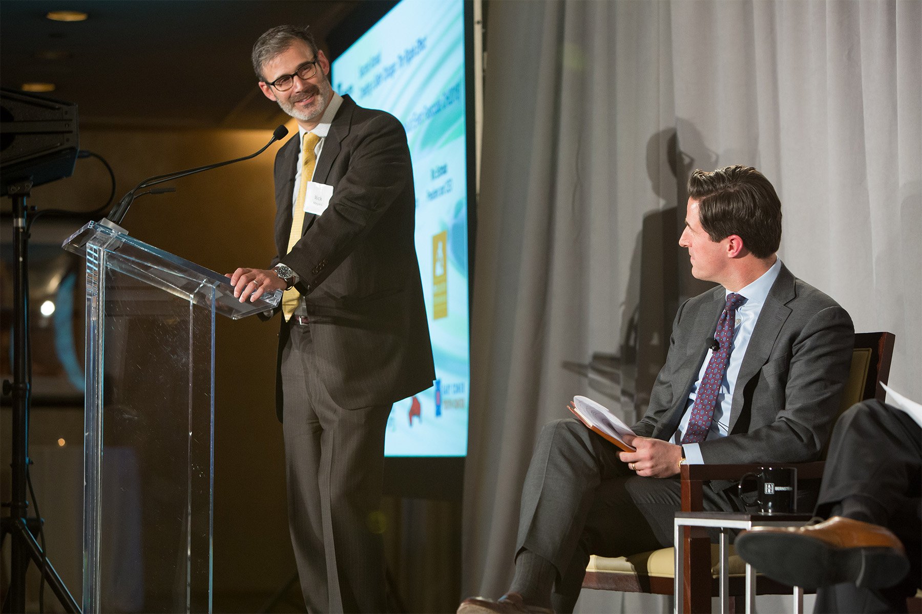 A man with glasses, gray hair, and a beard stands at a clear podium speaking into a microphone, wearing a dark suit, white shirt, and yellow tie. Another man with dark hair, dressed in a gray suit and patterned tie, sits to the right, holding an open