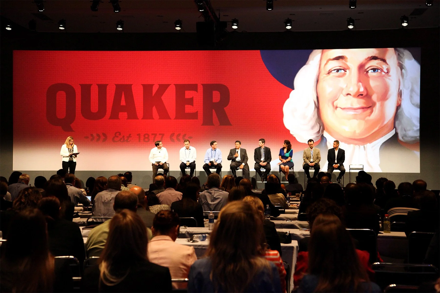 A large audience watching a panel discussion on stage with nine people seated in front of a giant screen displaying the Quaker logo and a portrait of a woman with white hair. A woman stands to the left, reading from a tablet, and a man is speaking in