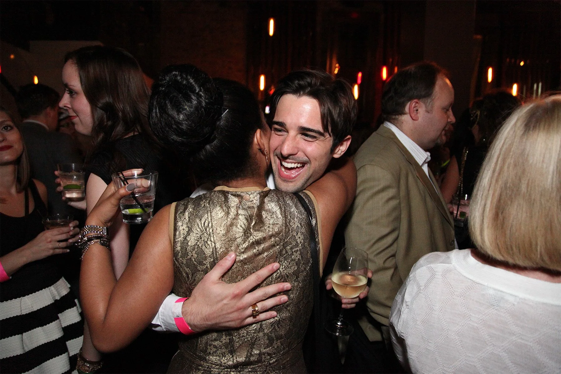 People at a social gathering hugging and holding drinks in a dimly lit setting, likely a party or celebration.